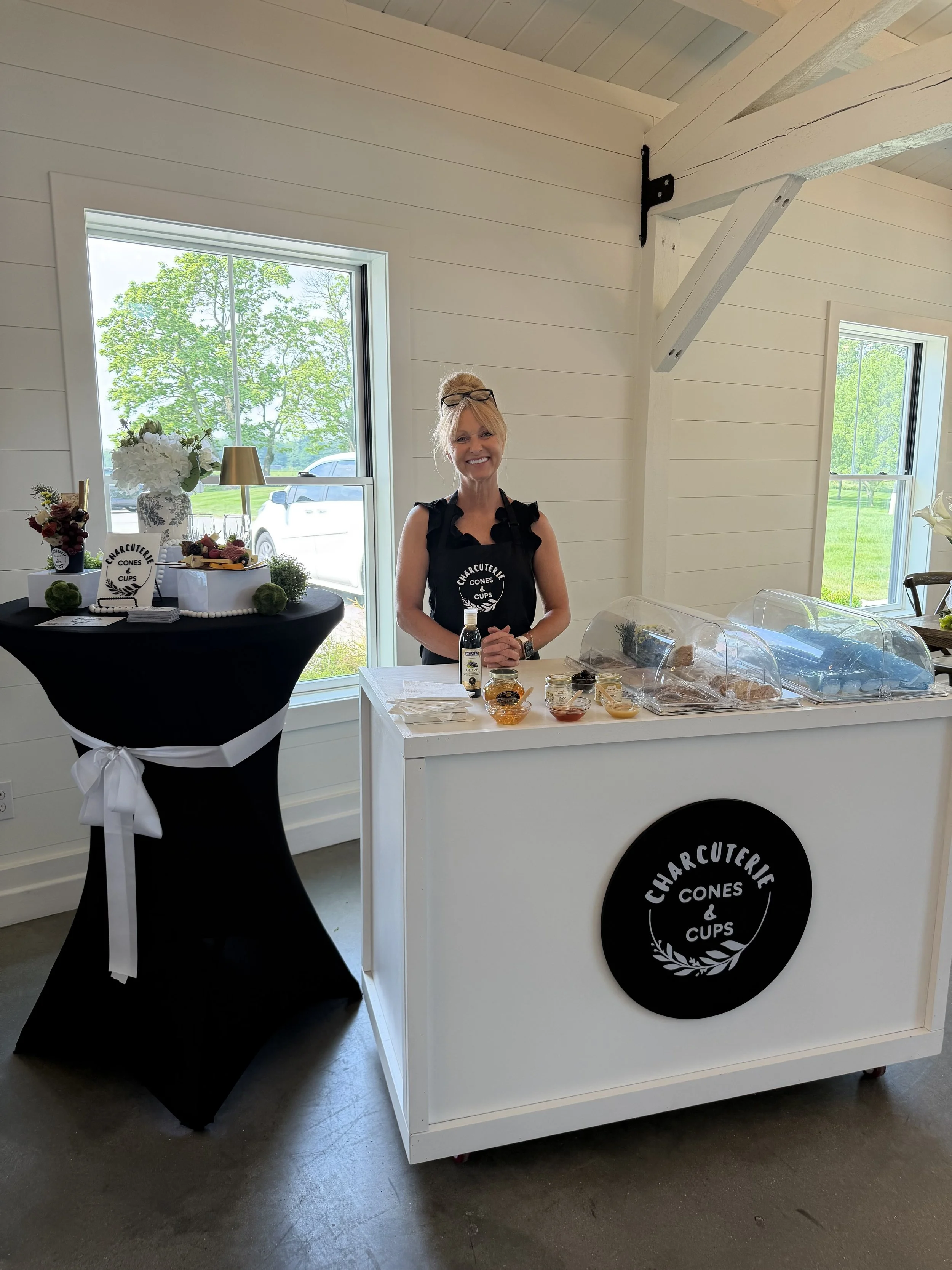 A smiling woman standing behind a white food cart with a black circular sign that reads 'Charcuterie Cones & Cups'. The cart has a clear top displaying food items, and there are small bowls with condiments on the white surface. To her left on a black