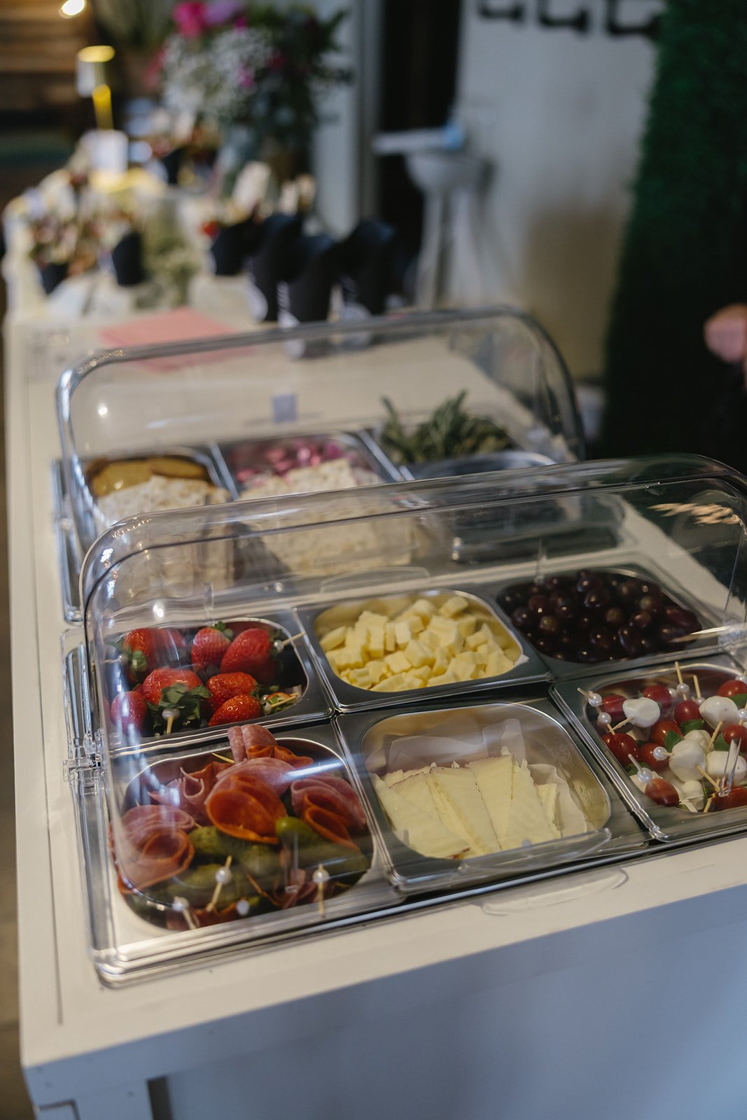 A spread of cheese, strawberries, grapes, and meats at a buffet station with a clear glass cover.