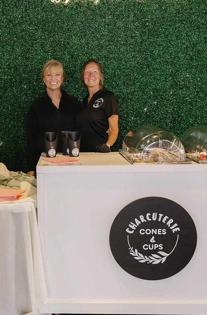 Two women standing behind a white counter at a charcuterie stand with a black circular sign that reads "Charcuterie Cones & Cups". They are smiling, one dressed in black and the other in a black shirt with a logo. The background is a green wall made 