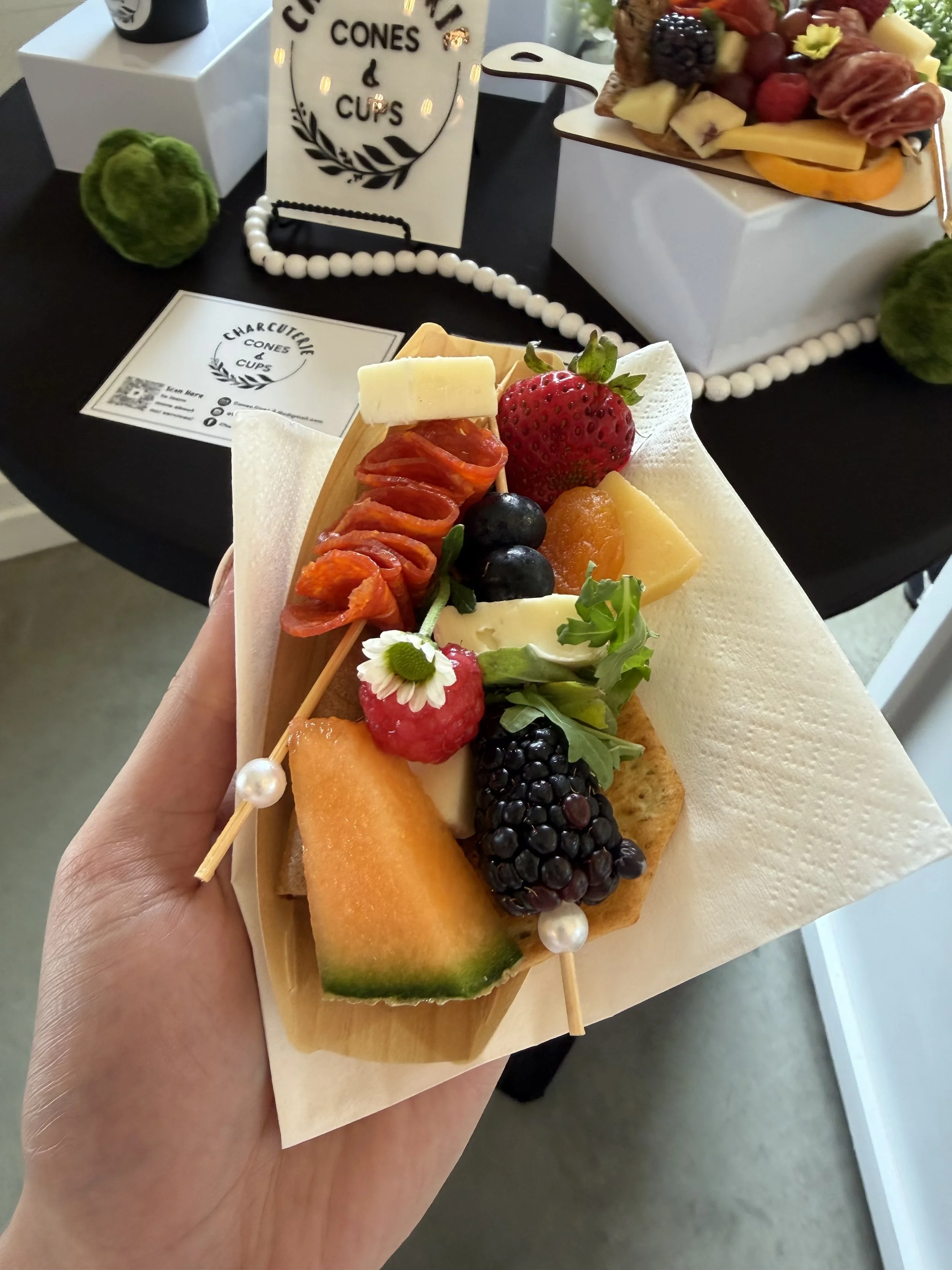 A hand holding a wooden platter with various fresh fruits, including a strawberry, blueberries, blackberries, raspberry, chunks of cantaloupe and honeydew, a piece of white cheese, and a small edible flower, wrapped in a white paper towel.