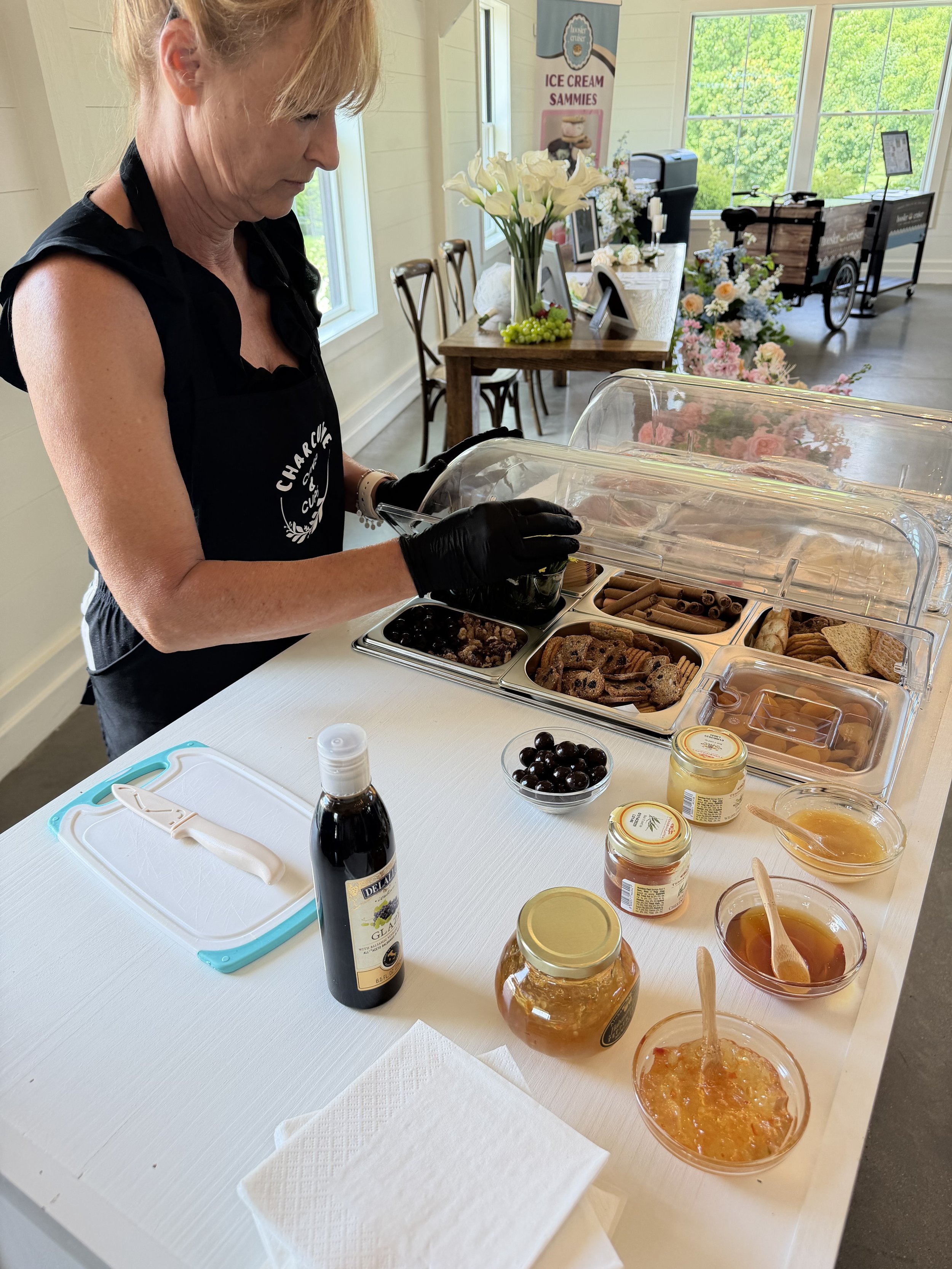 A woman wearing a black apron and gloves is serving ice cream from a countertop with various containers of toppings and sauces. In the background, there are tables with floral arrangements, a flag/banner, and windows showing green trees outside.