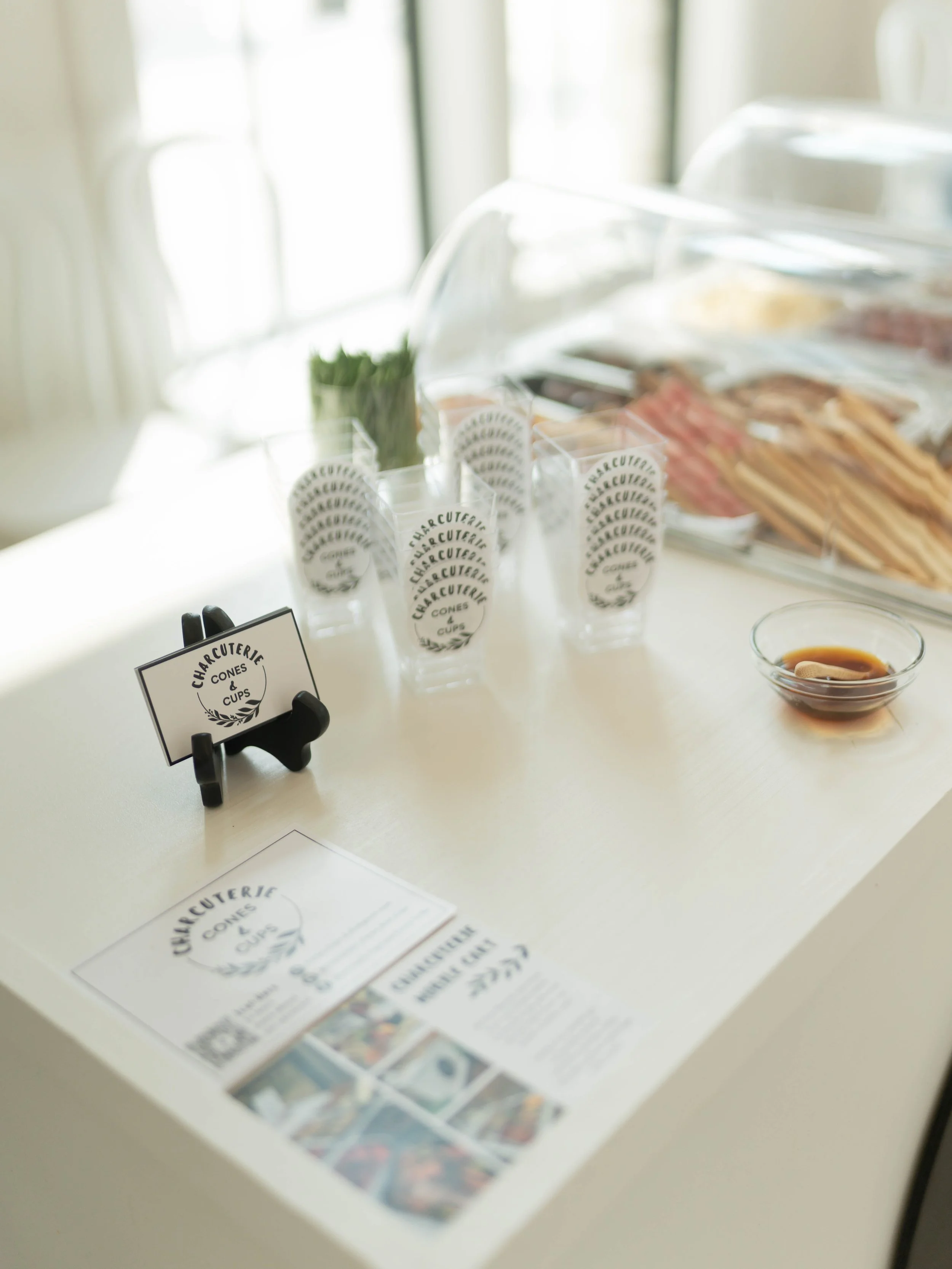 Clear plastic containers holding bacon, wrapped cheese, and herbs on a white countertop with a sign reading 'Charcuterie Cones & Cups' and a small dish of sauce.
