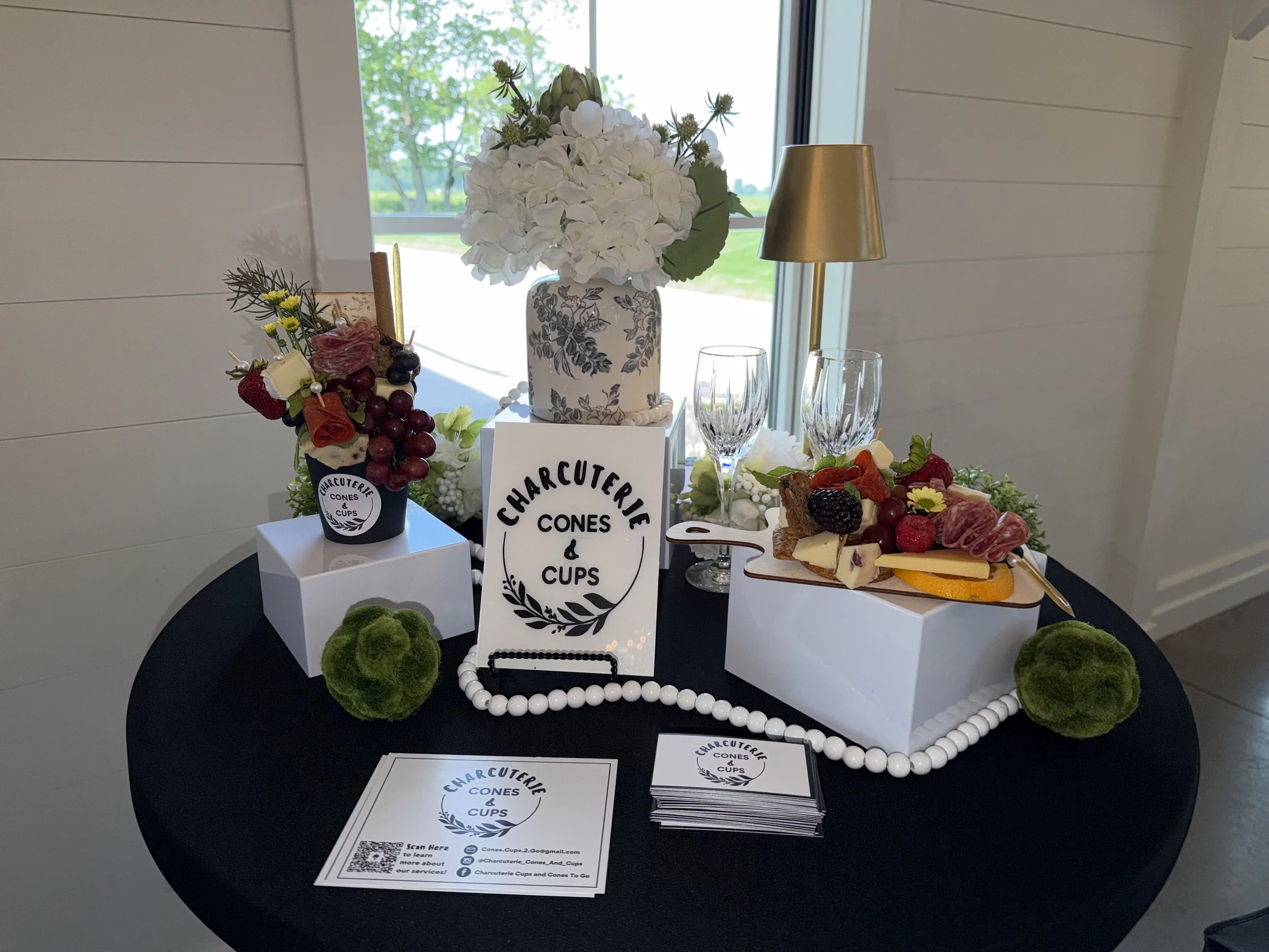 Display of charcuterie cones and cups on a black table, with a white vase of white flowers, a golden lamp, and a sign reading 'Charcuterie Cones & Cups' in front of a window with trees outside.
