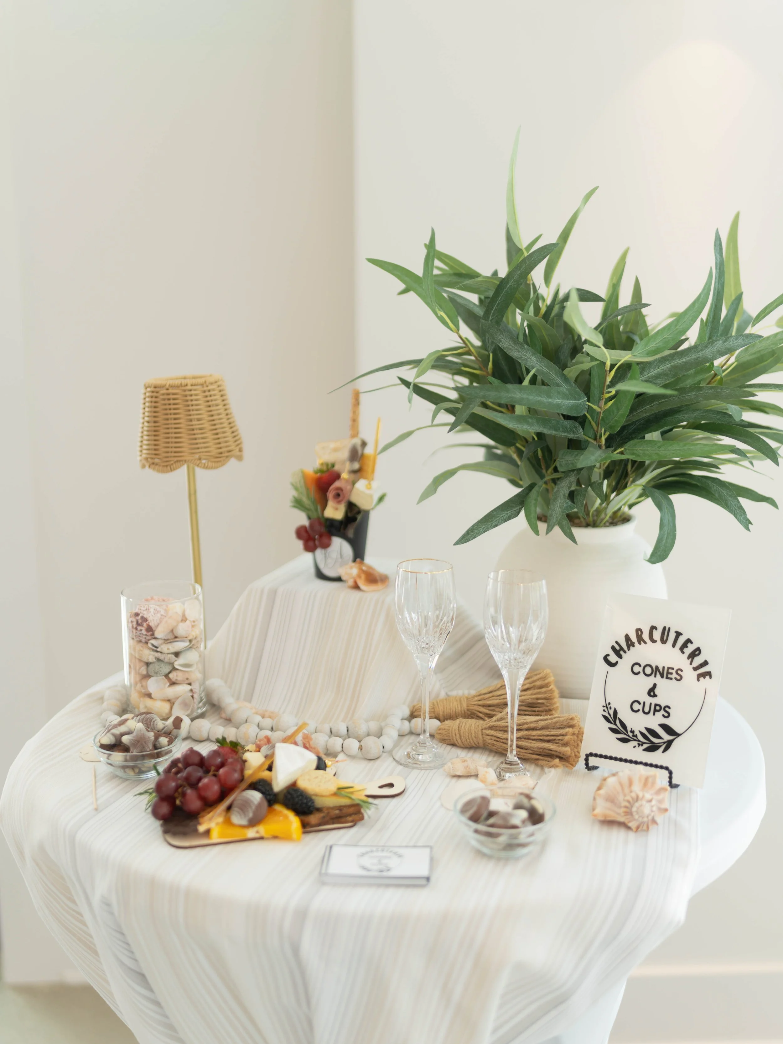 Table decorated with beach-themed items including seashells, grapes, desserts, and a sign that says "Charcuterie Cones & Cups." There is a large green plant in a white pot, a small wicker lamp, and a display of seashells and stones arranged on the ta