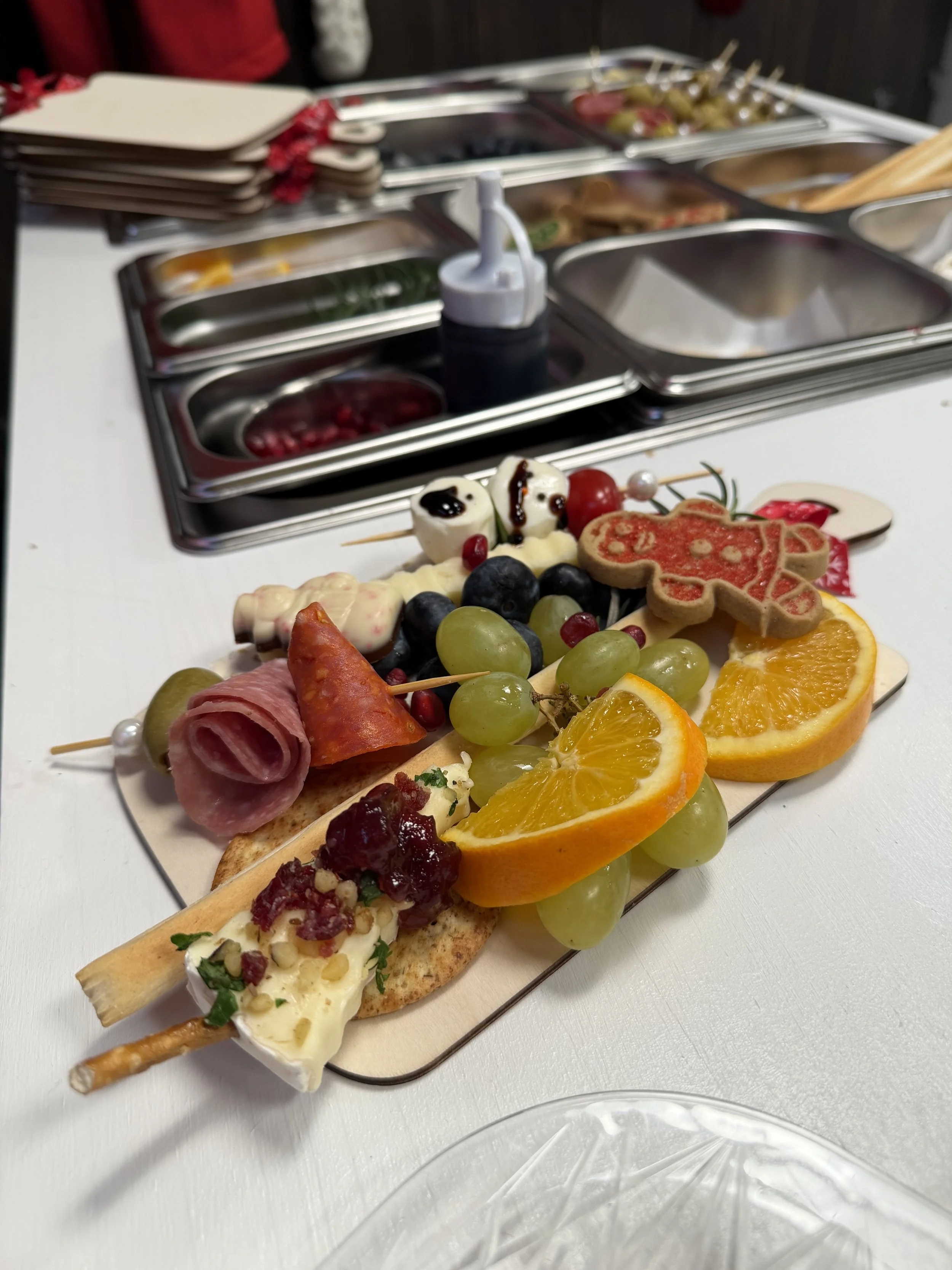 Close-up of a tray with assorted appetizers including grapes, oranges, cheese, meats, and a breadstick, with a blurred buffet setup in the background.