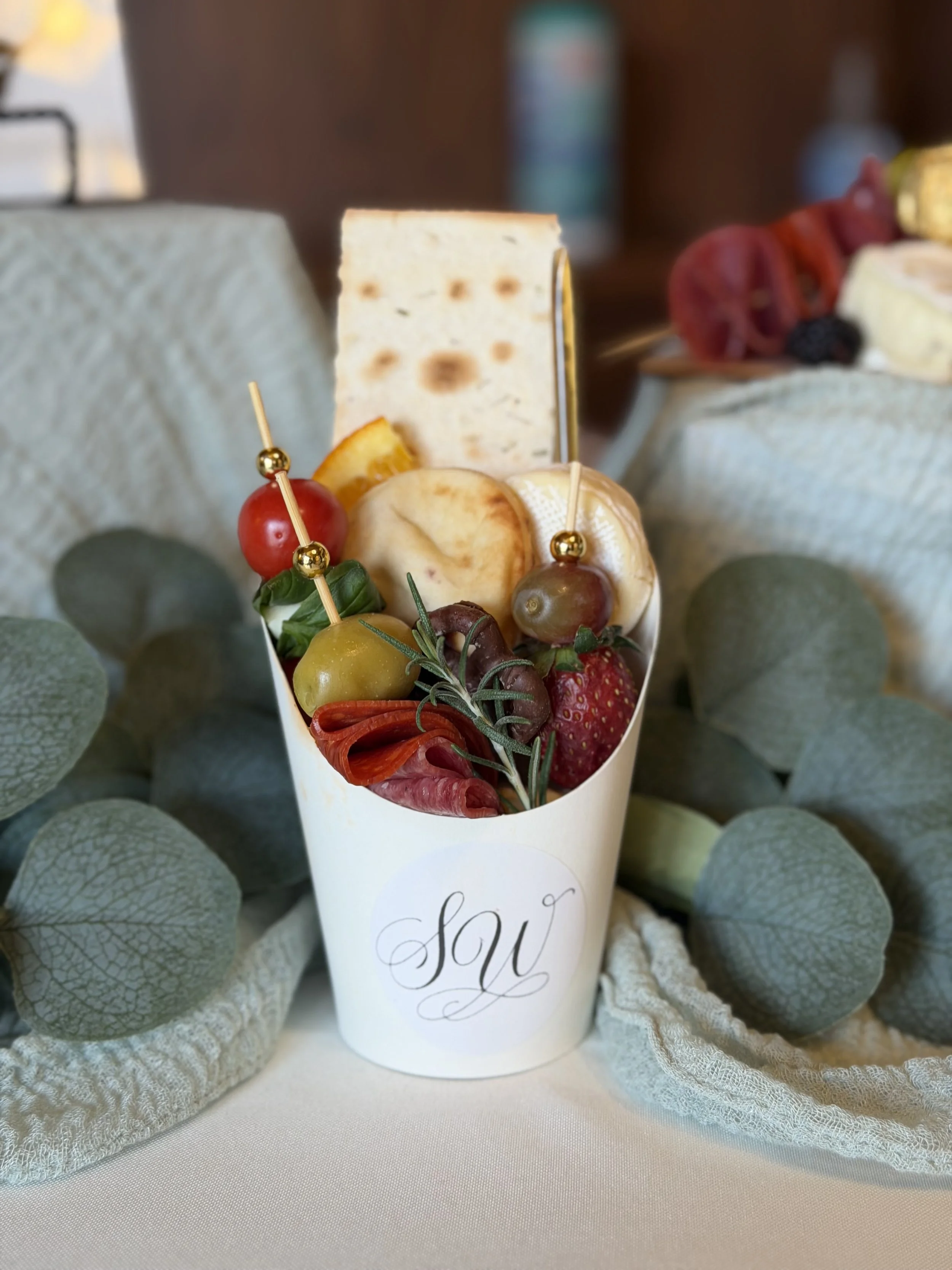 A decorative arrangement of cheese, fruits, and berries in a cup with the word 'you' written on it, surrounded by a soft fabric and eucalyptus leaves.