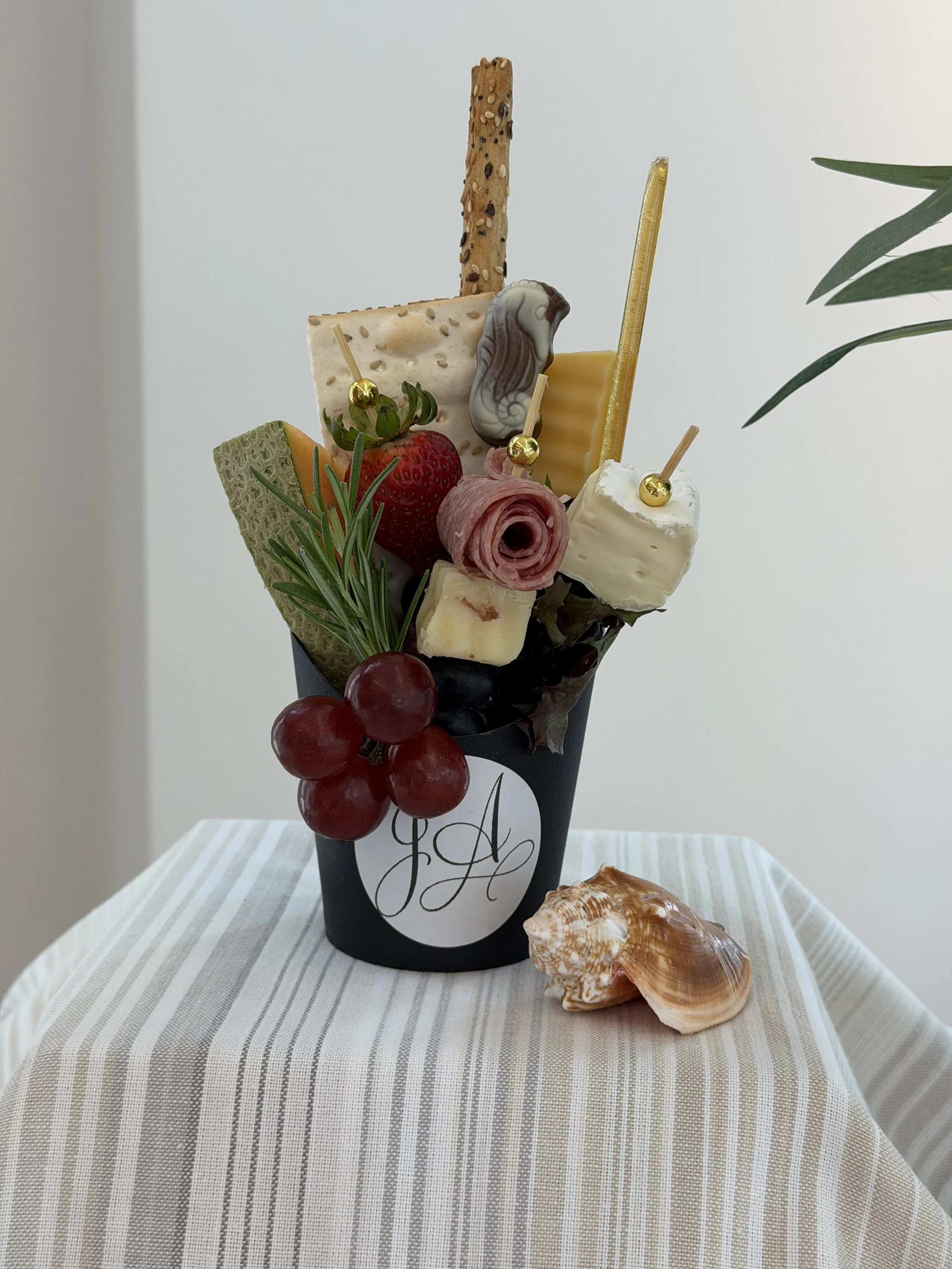 A bouquet of assorted cheeses, grapes, strawberries, a sprig of rosemary, and decorative crackers in a black cone-shaped container with monogrammed initials, placed on a striped tablecloth with seashells nearby.