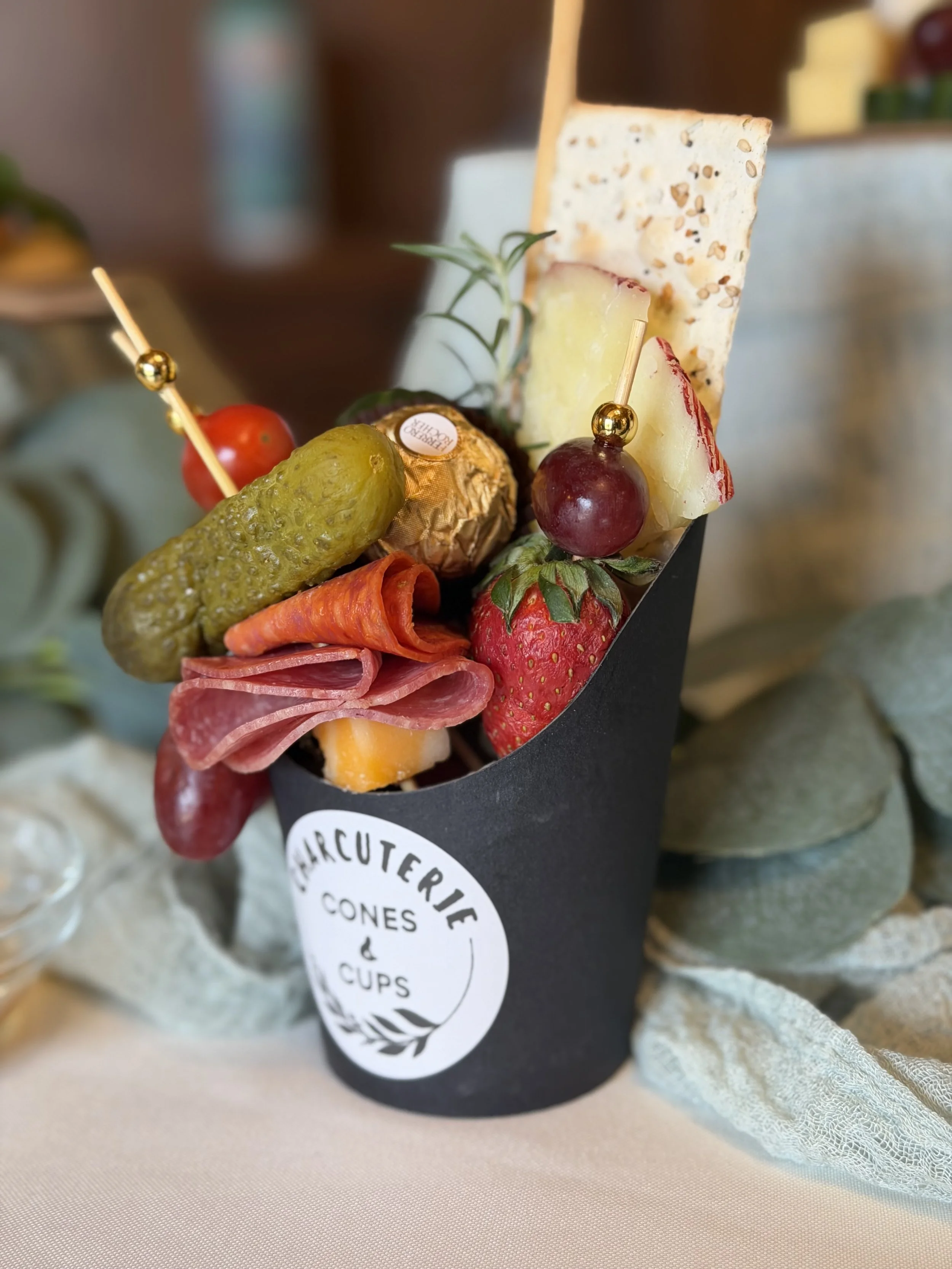 An assortment of cheeses, strawberries, grapes, cherry tomato, and cured meats presented in a black cone-shaped holder with a logo, placed on a light-colored surface with a textured cloth underneath.