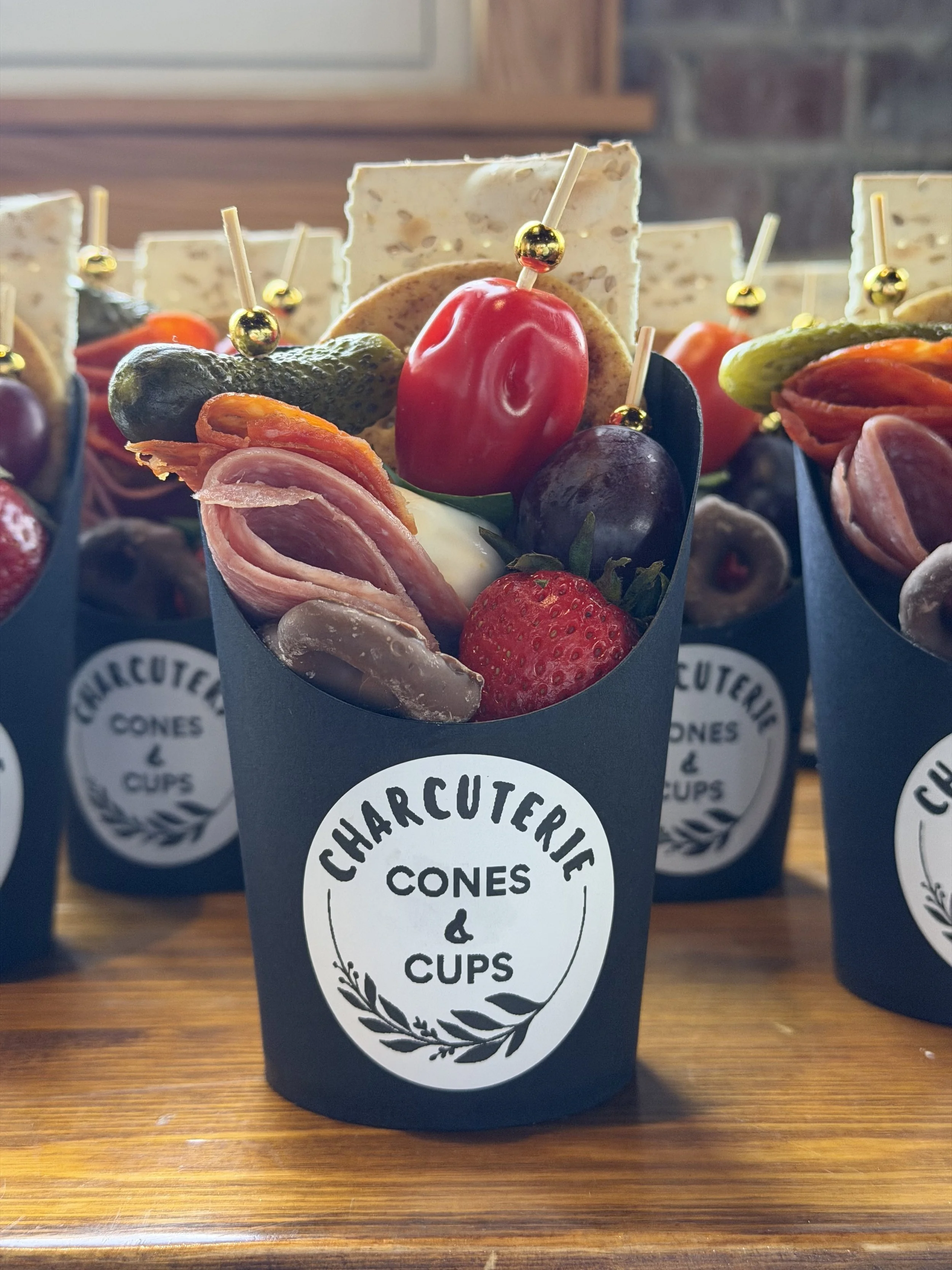 Various charcuterie items including slices of cured meats, cheese, grapes, strawberries, pickles, crackers, and pepper in a paper cone with a label that reads 'Charcuterie Cones & Cups'.