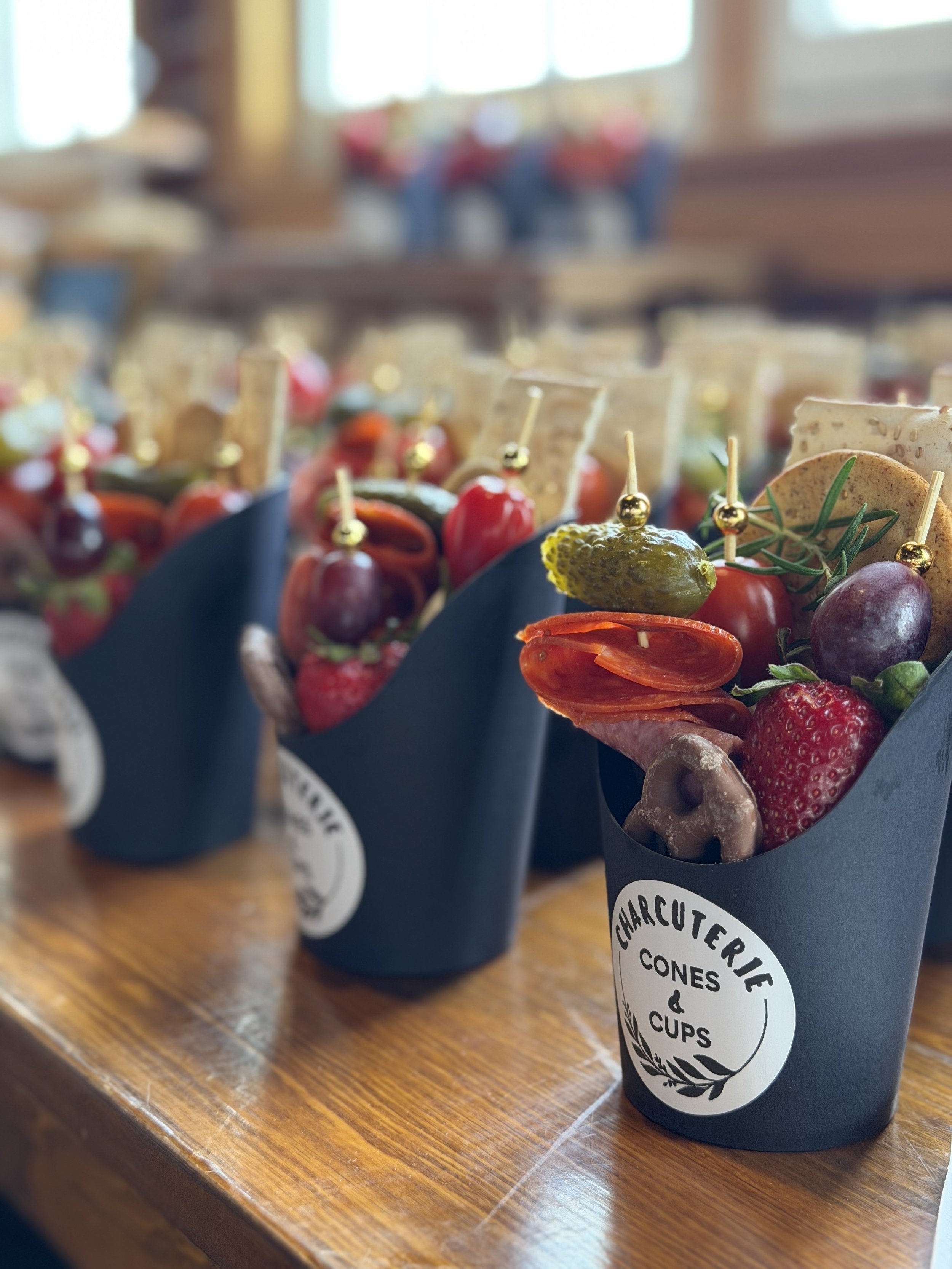 A black paper cone with a label reading 'Charcuterie Cones & Cups' containing various snacks including grapes, cherry tomatoes, olives, bread slices, and cheese, on a wooden table with similar cones in the background.