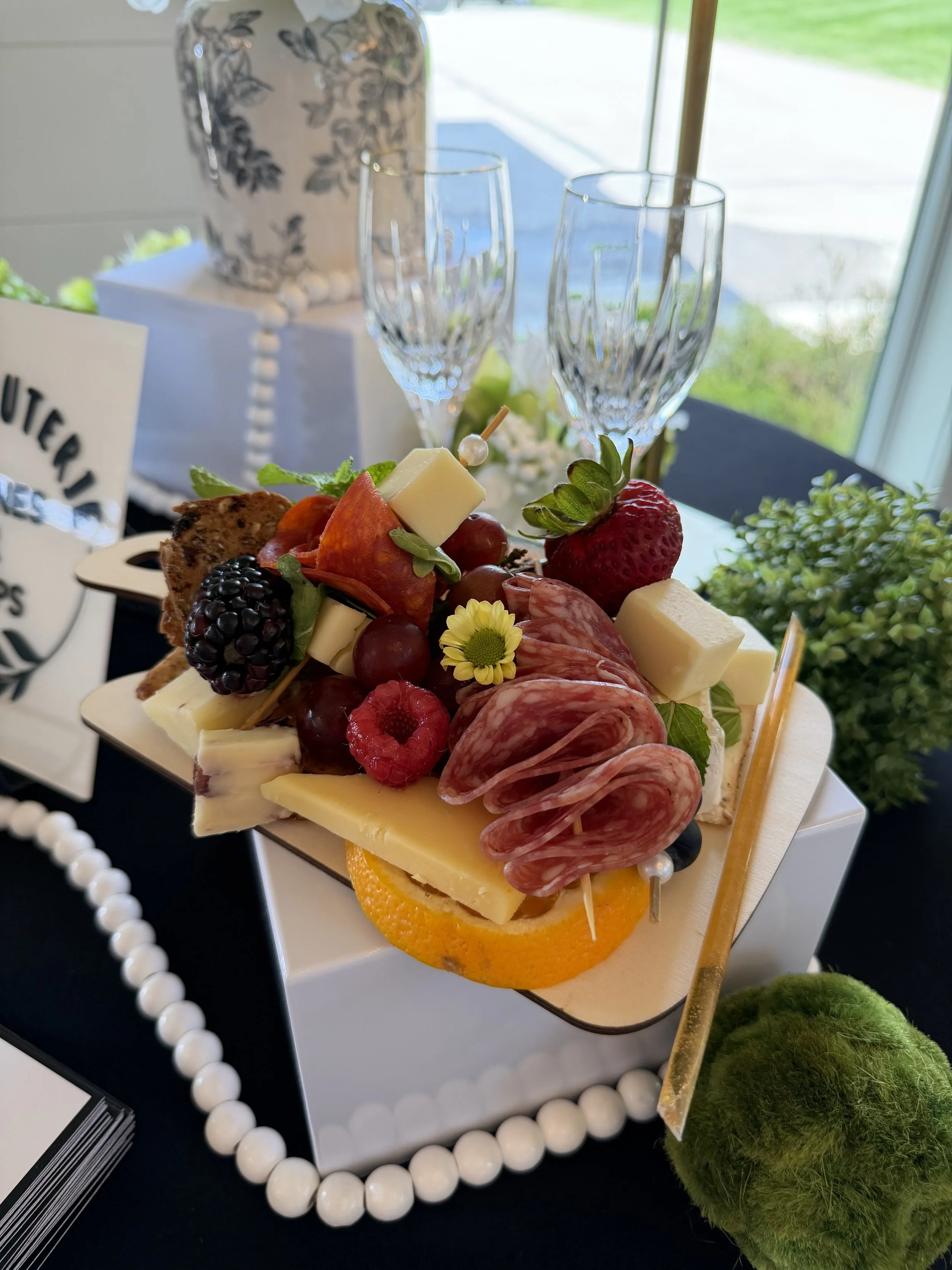 A platter of assorted cheeses, meats, grapes, strawberries, blackberries, and vegetables arranged on a board, with a decorative flower and two empty champagne glasses in the background.