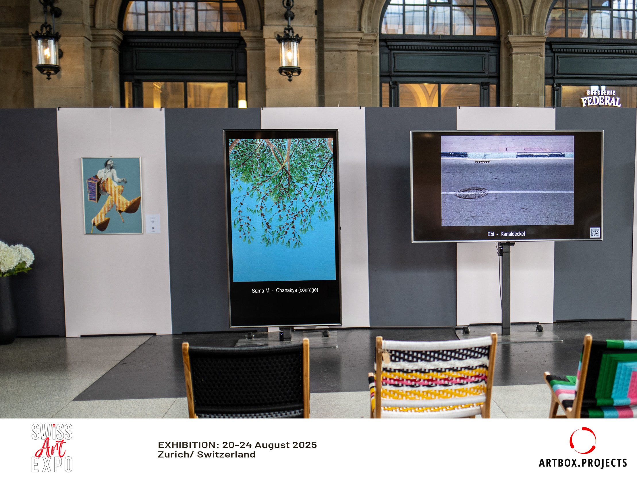 Art exhibition display with  framed artwork on a dark and light grey board at Zurich Switzerland Swiss Art Expo 2025, held in a historic building with arched windows and vintage hanging lanterns, with chairs in front of the display.