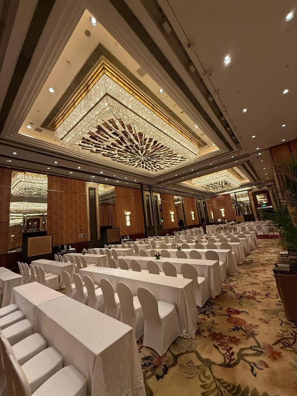 Elegant banquet hall with white tablecloth-covered tables and chairs, ornate chandeliers on the ceiling, and decorative wall panels.