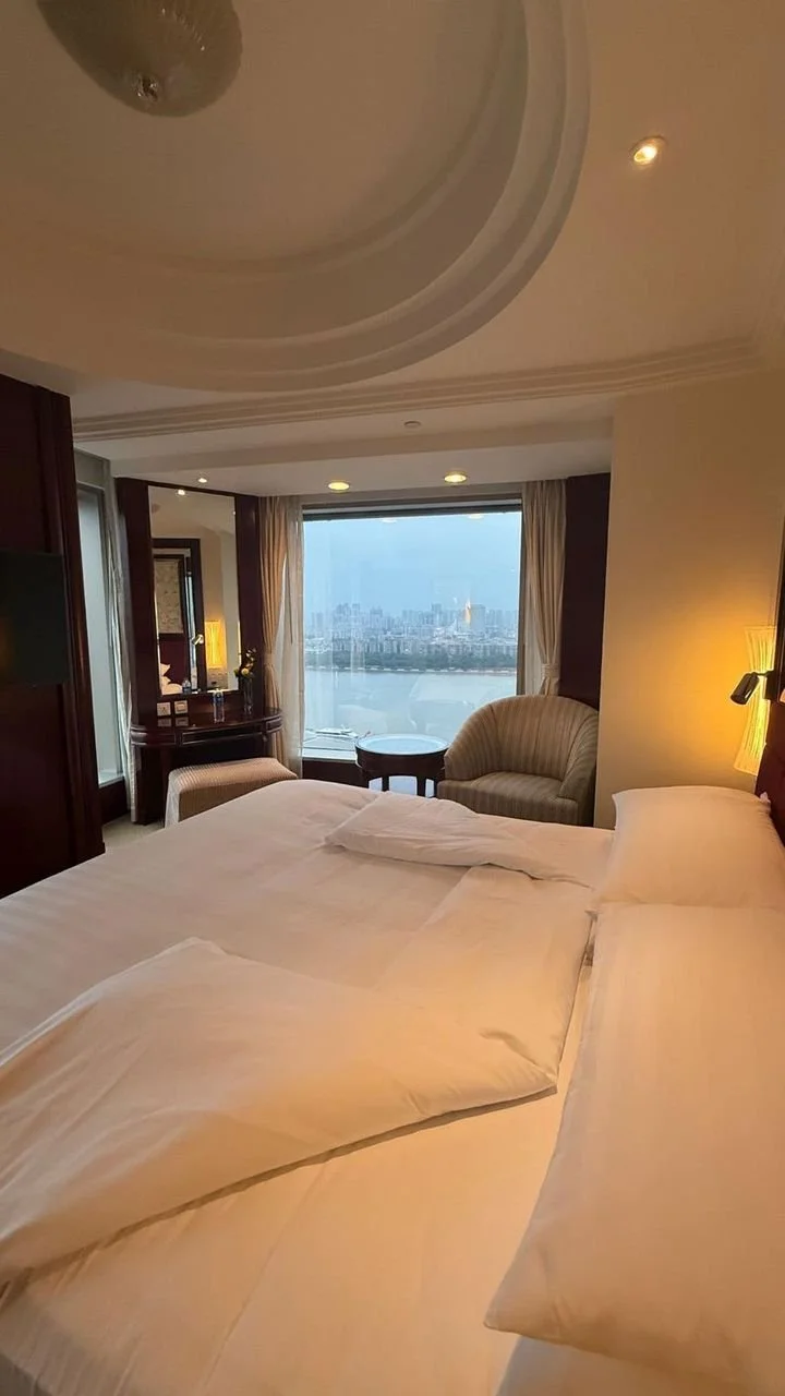 Hotel room with a large bed, a window with cityscape view, and a sitting area with a chair and small table.