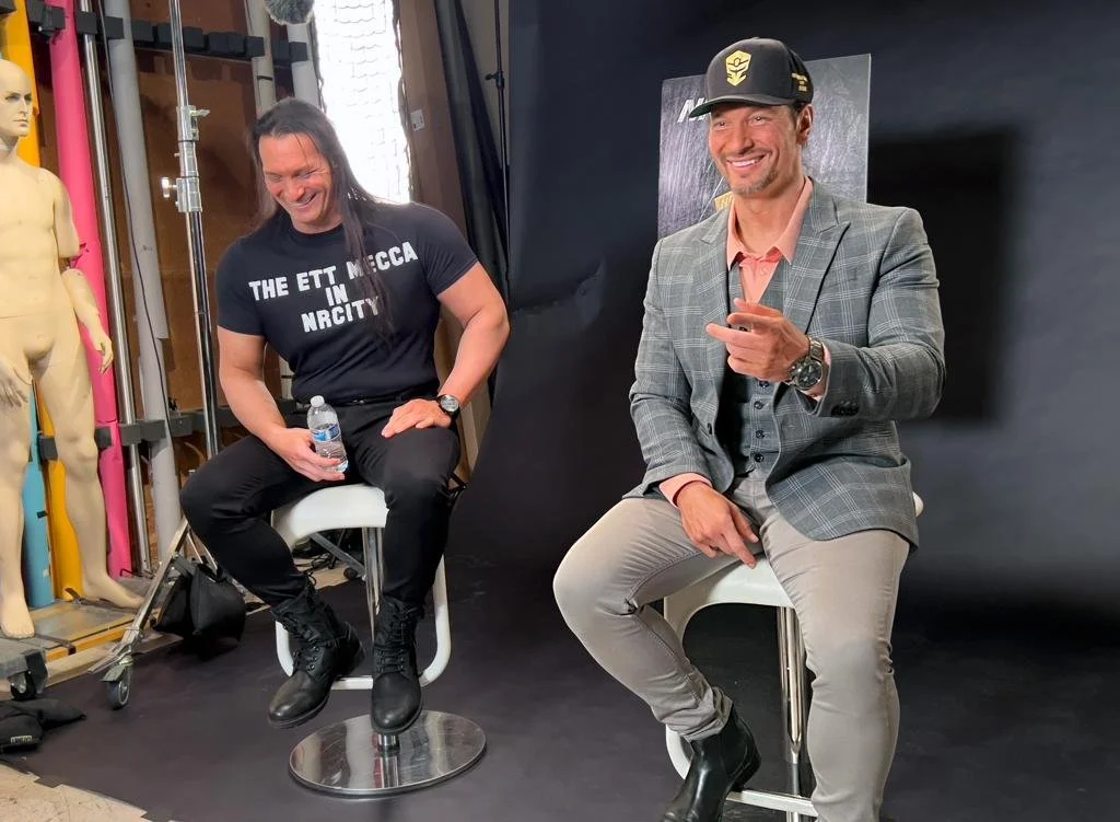 Two men sitting on stools in front of a black backdrop, smiling and laughing. One holds a water bottle, the other points towards the camera. MRI mannequins and medical equipment are visible on the left.