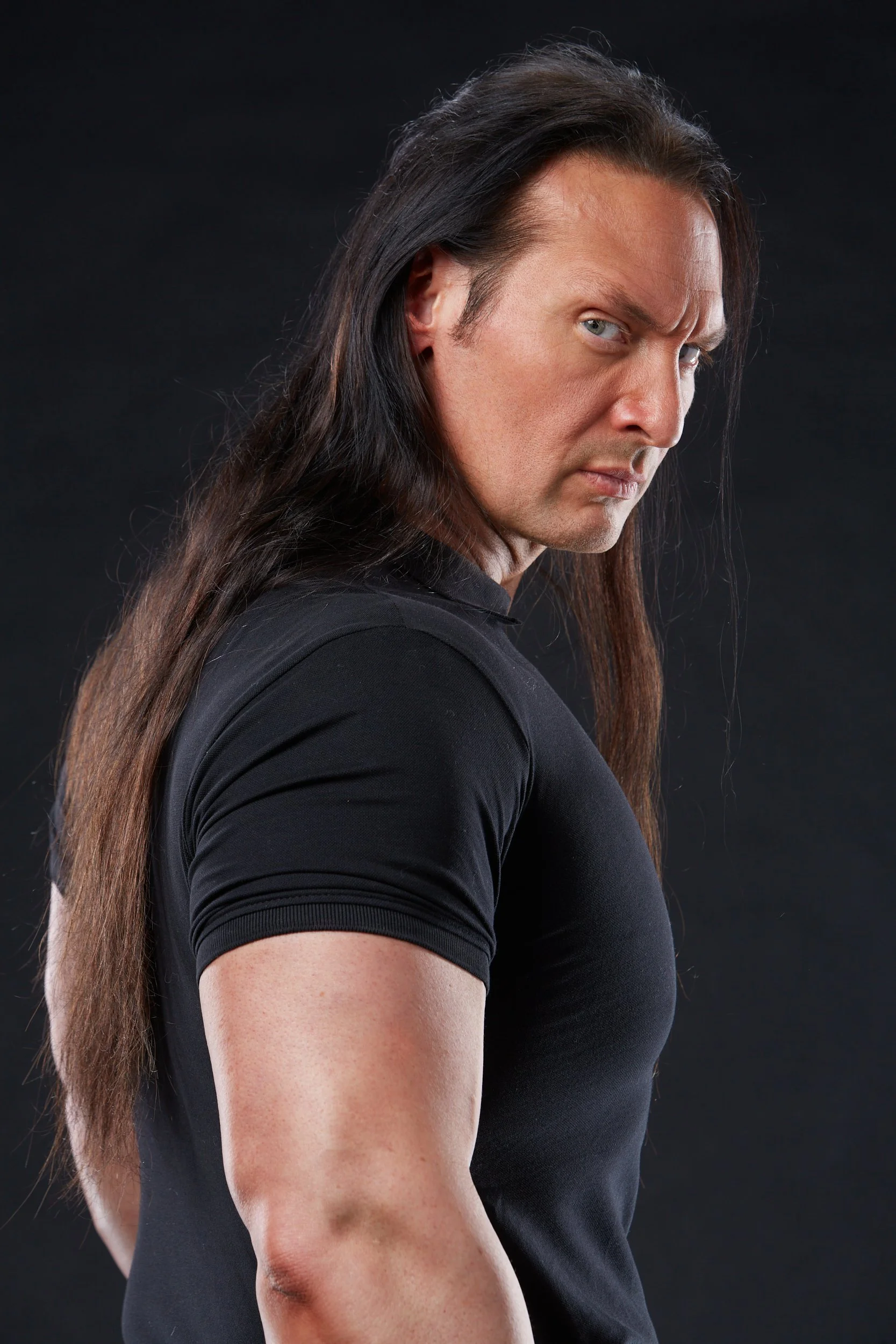 A man with long dark hair and a serious expression, wearing a black t-shirt, against a black background.