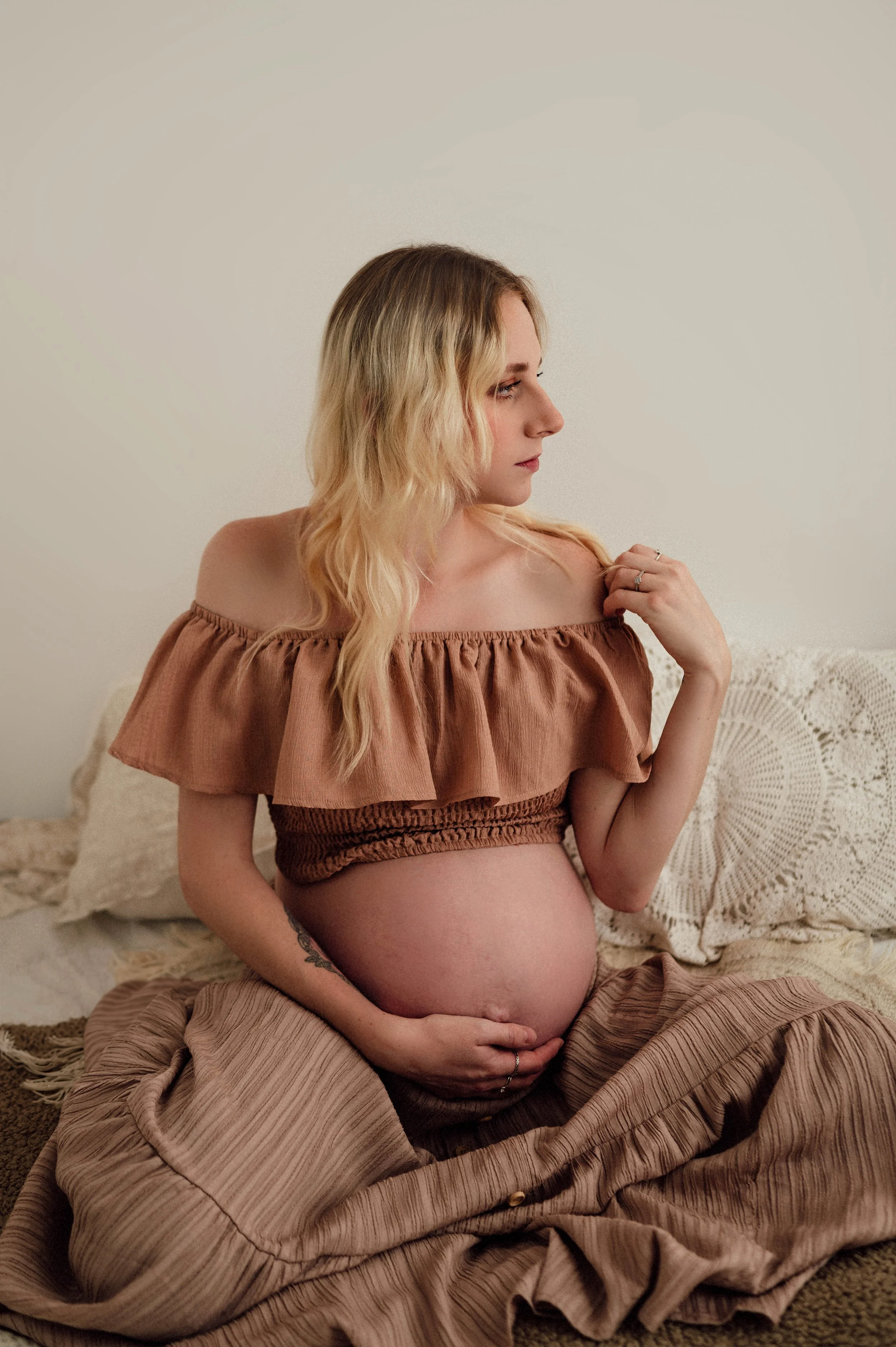 Mother holding her baby bump, sat on a bed with a two piece, crop top and skirt combo in pink and brown tones. Photography in wiltshire