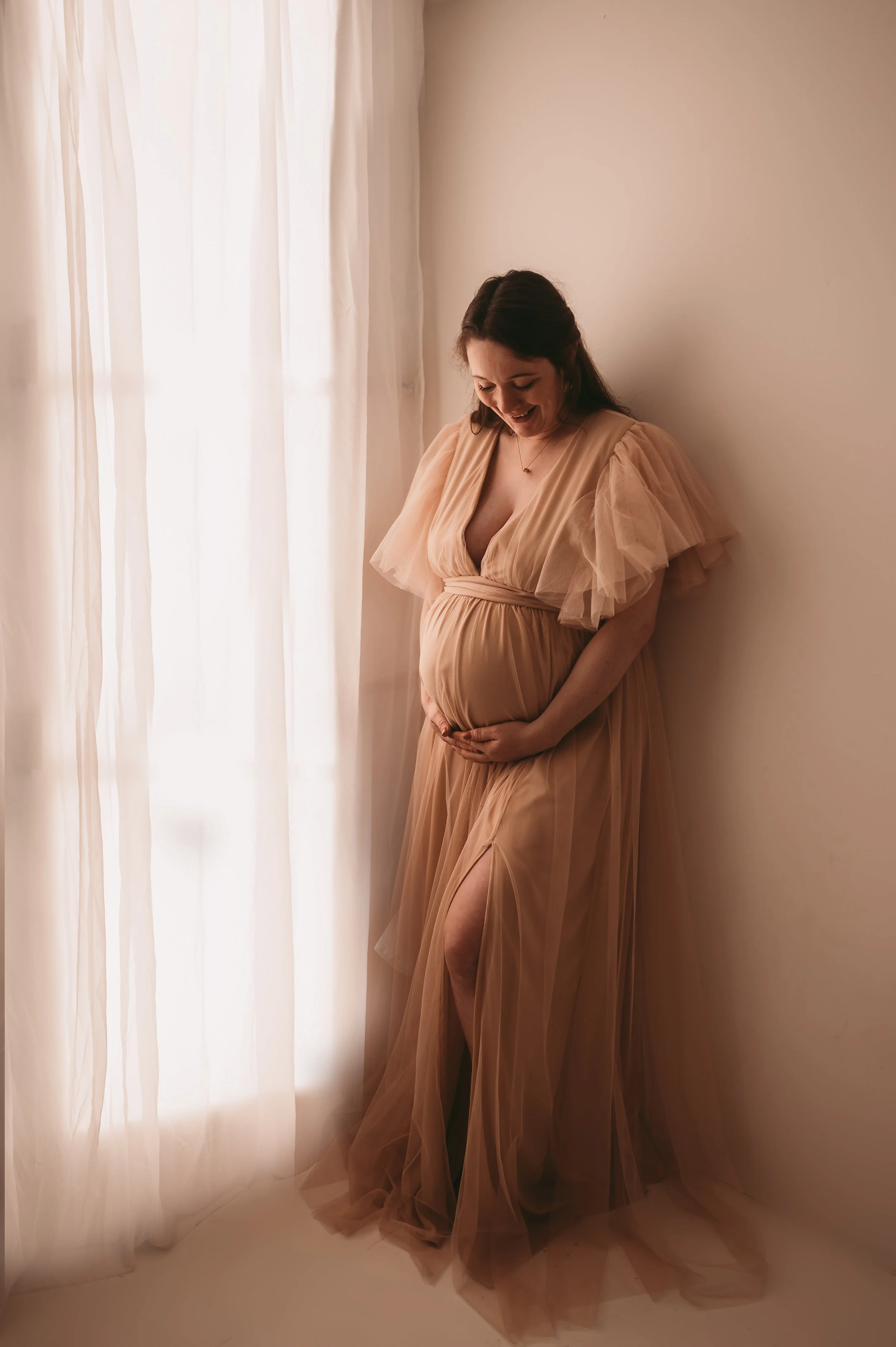 Maternity photograph in a photo studio next to a window, woman holding baby bump wearing a tulle floor length dress, Studio is in Trowbridge, Wiltshire