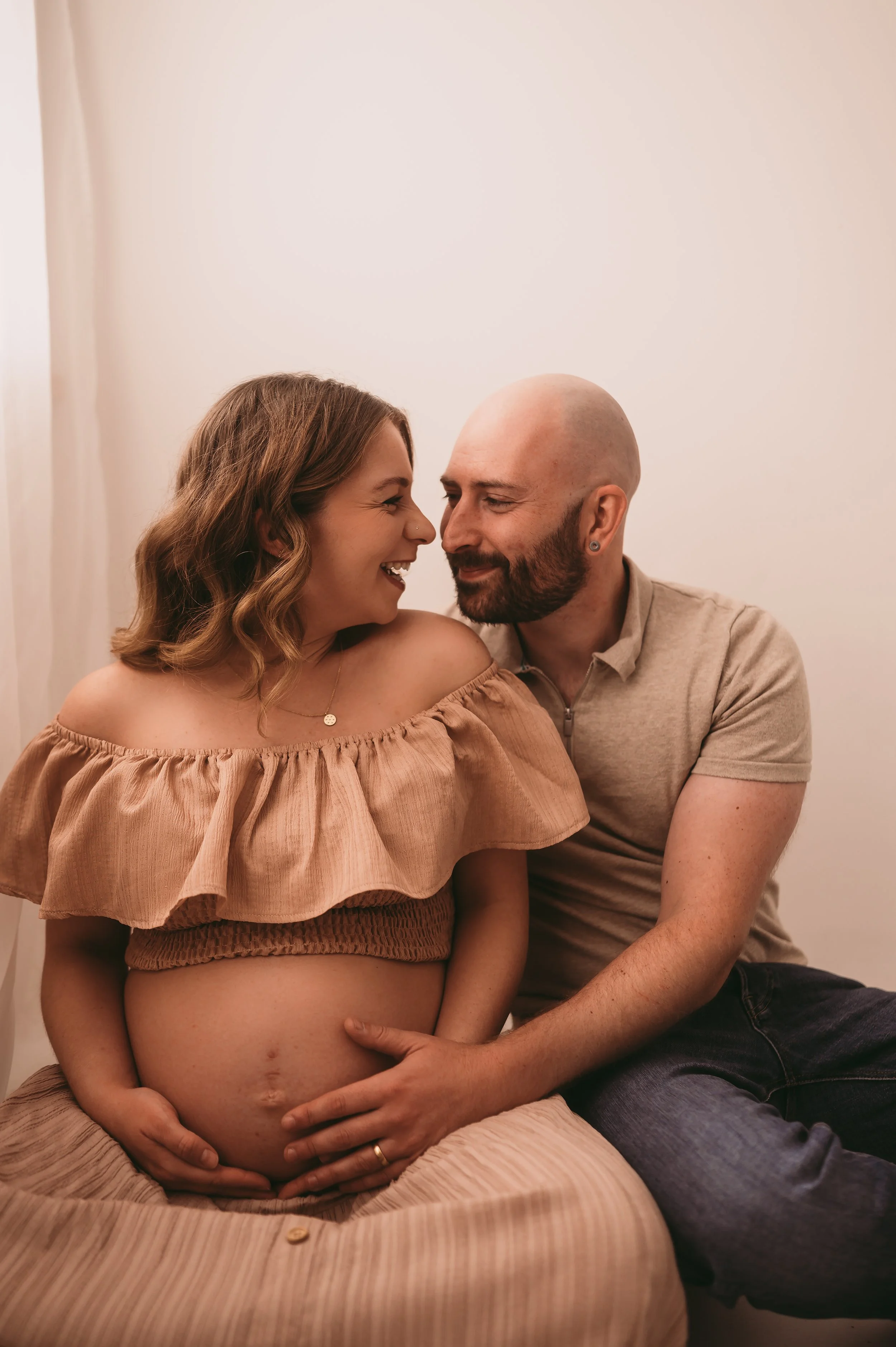 Maternity couples photographed in a studio in Wiltshire, the father is cuddling and holding baby bump whilst they look and smile at each other