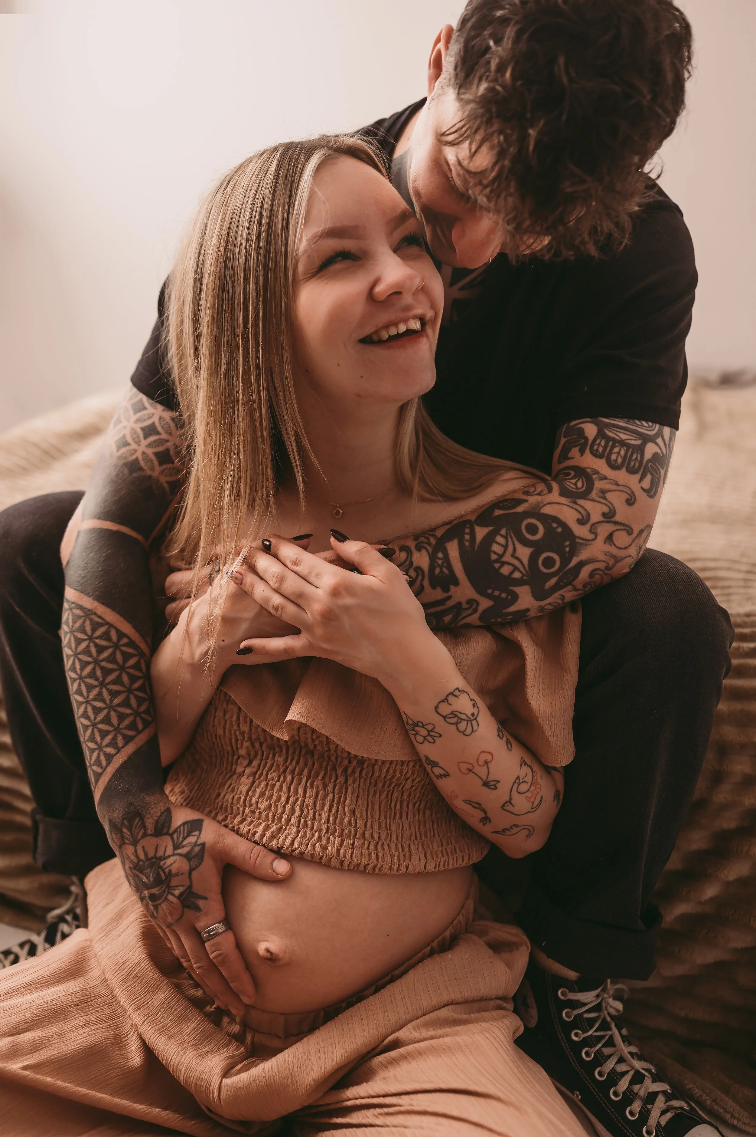 Couple cuddling each other and baby bump, sat on a bed and on the floor. The couple have tattoos and are smiling. Photographer is in Trowbridge, Wiltshire
