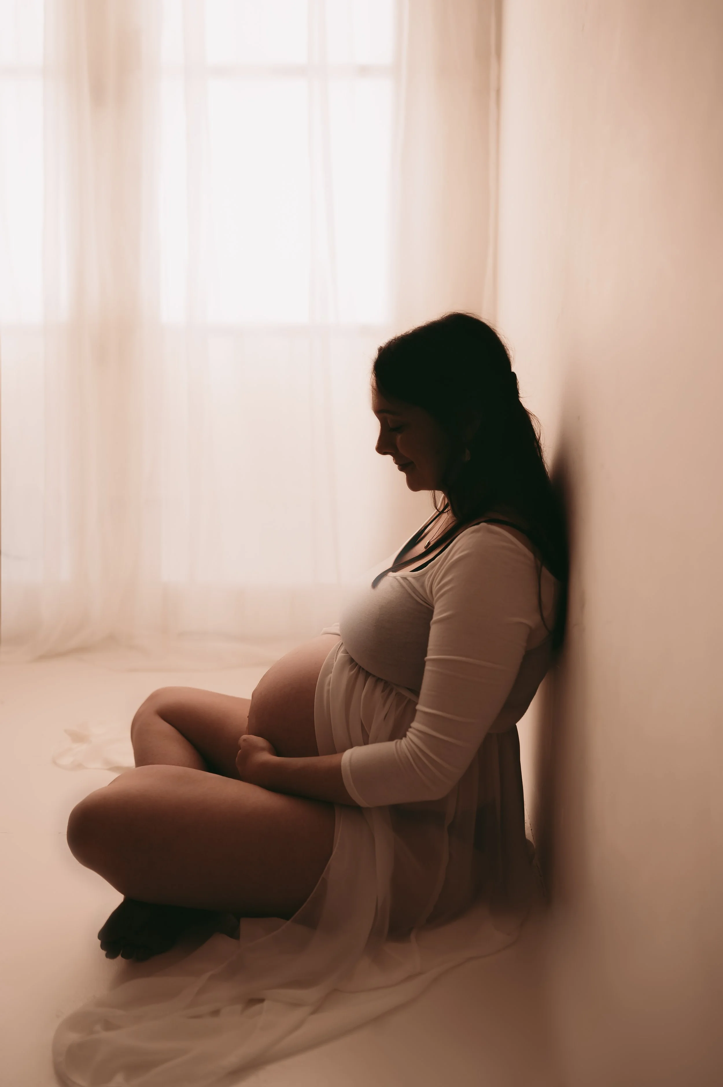 A Woman sat on the floor, window in the background and her and baby bump in a silhouette,  Maternity photography by kp-photo, based in Wiltshire