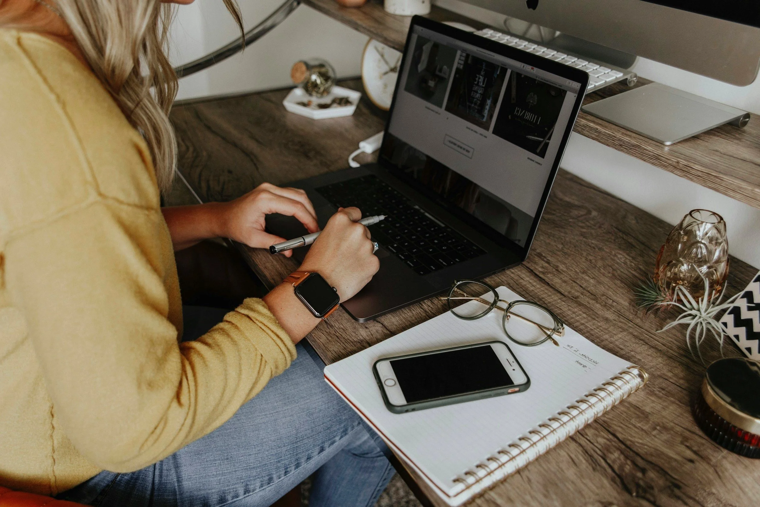 A person sitting at a wooden desk using a laptop, with a smartphone, glasses, notebook, and decorative items on the desk.