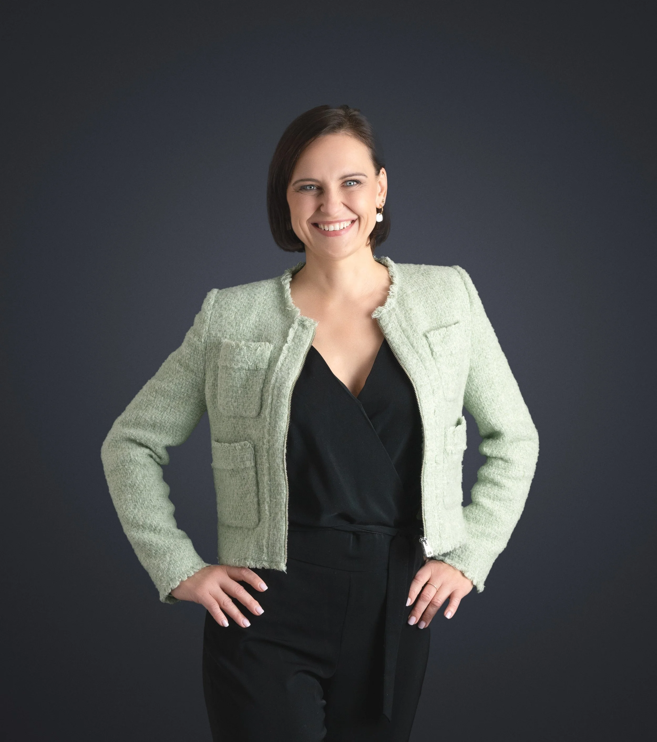 Lächelnde Frau in schwarzem Outfit mit grüner Jacke vor dunklem Hintergrund.