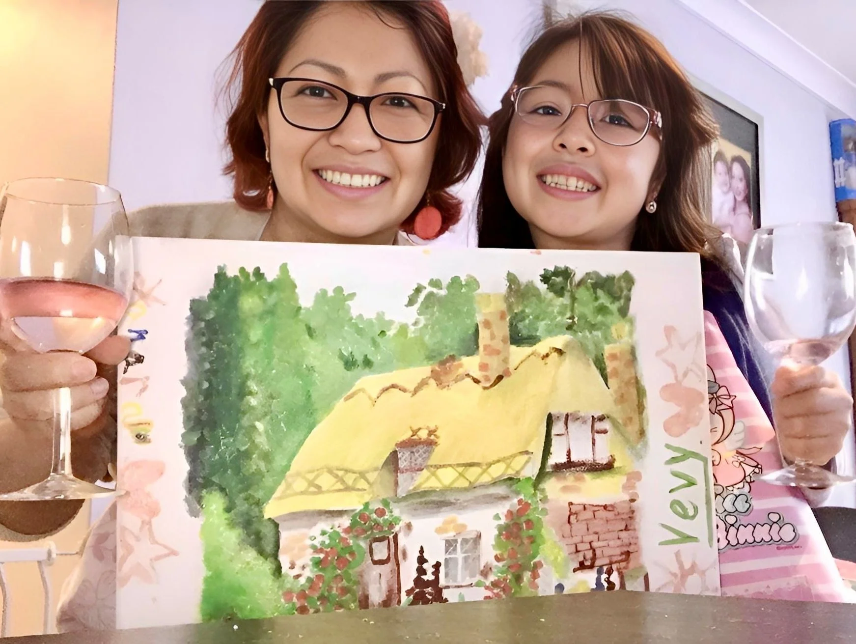 Two women smiling and holding wine glasses and a colorful drawing of a house with trees, flowers, and a brick fence, celebrating a special occasion.