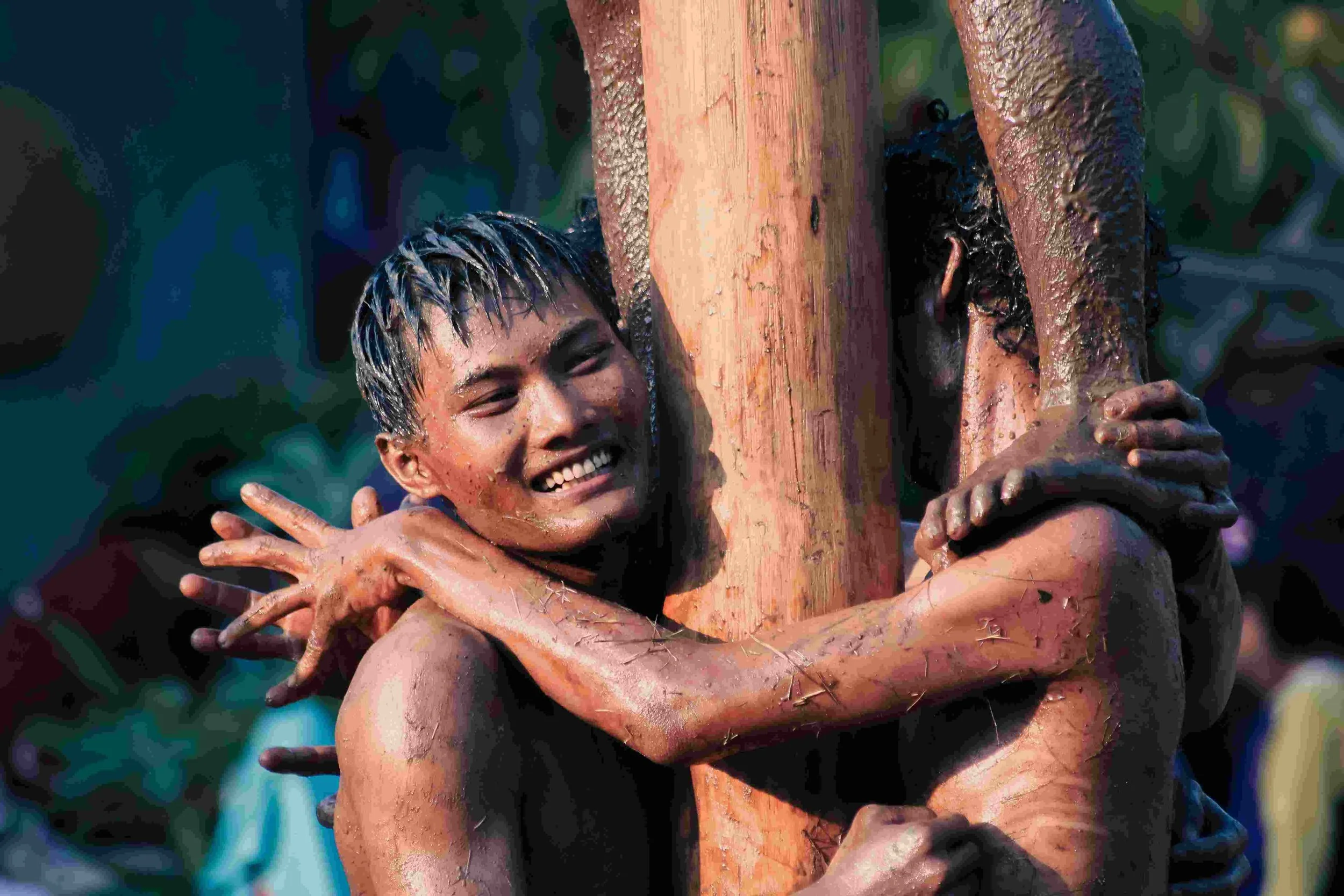 Two young men embracing each other around a wooden post, smiling and covered in mud, outdoors during daytime.