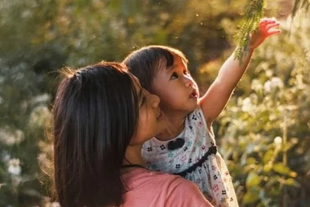 A woman holding a young girl outdoors, with sunlight shining through trees. The girl is reaching up and pointing at something in the sky.