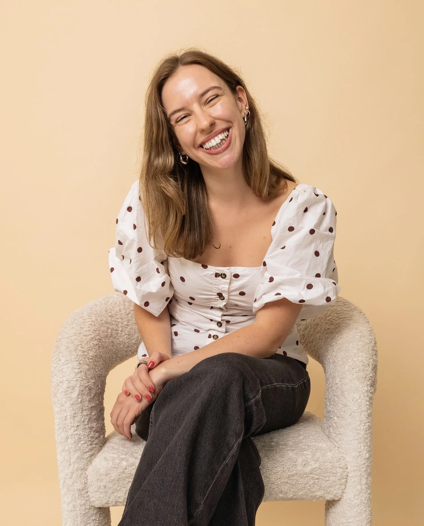 A woman with long brown hair, smiling and laughing, sitting on a plush beige chair against a pale peach background. She is wearing a white puff-sleeve blouse with brown polka dots and dark jeans.