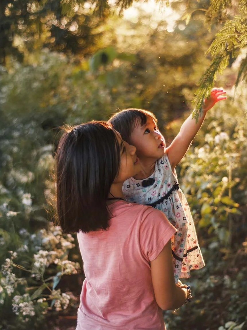 A woman holding a young girl outdoors, with the girl reaching up to touch tree branches. The scene is bathed in warm, golden sunlight, surrounded by greenery.