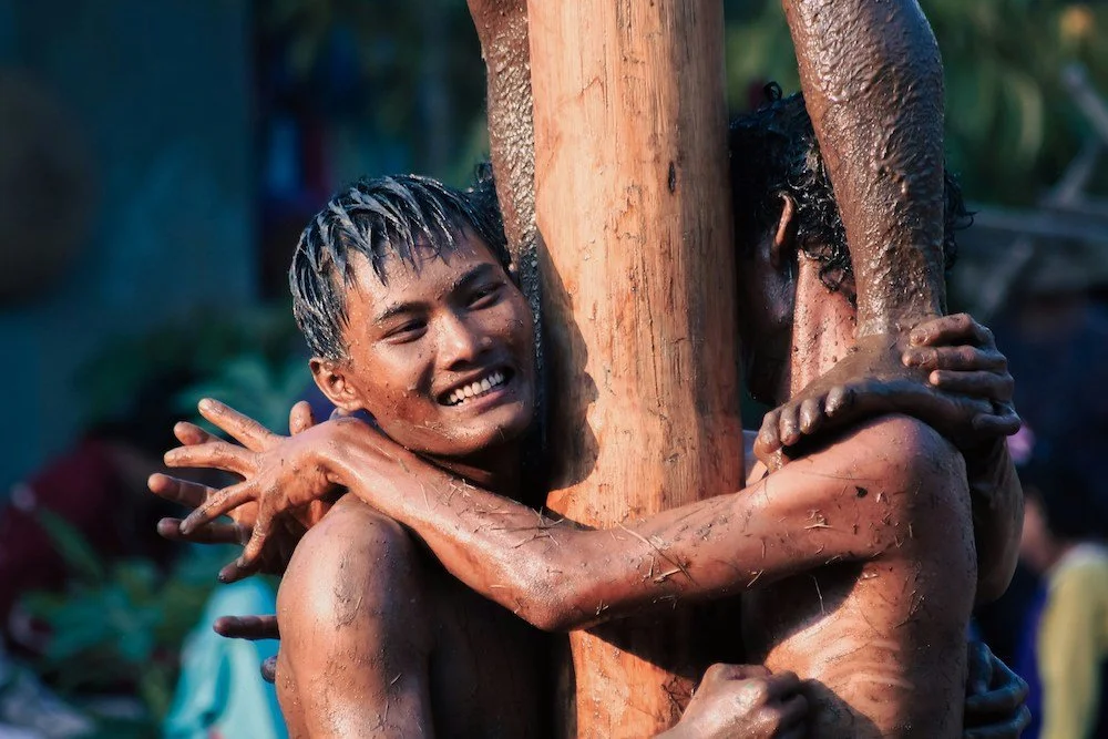 Two young men with wet hair and skin hugging each other around a wooden post in an outdoor setting.
