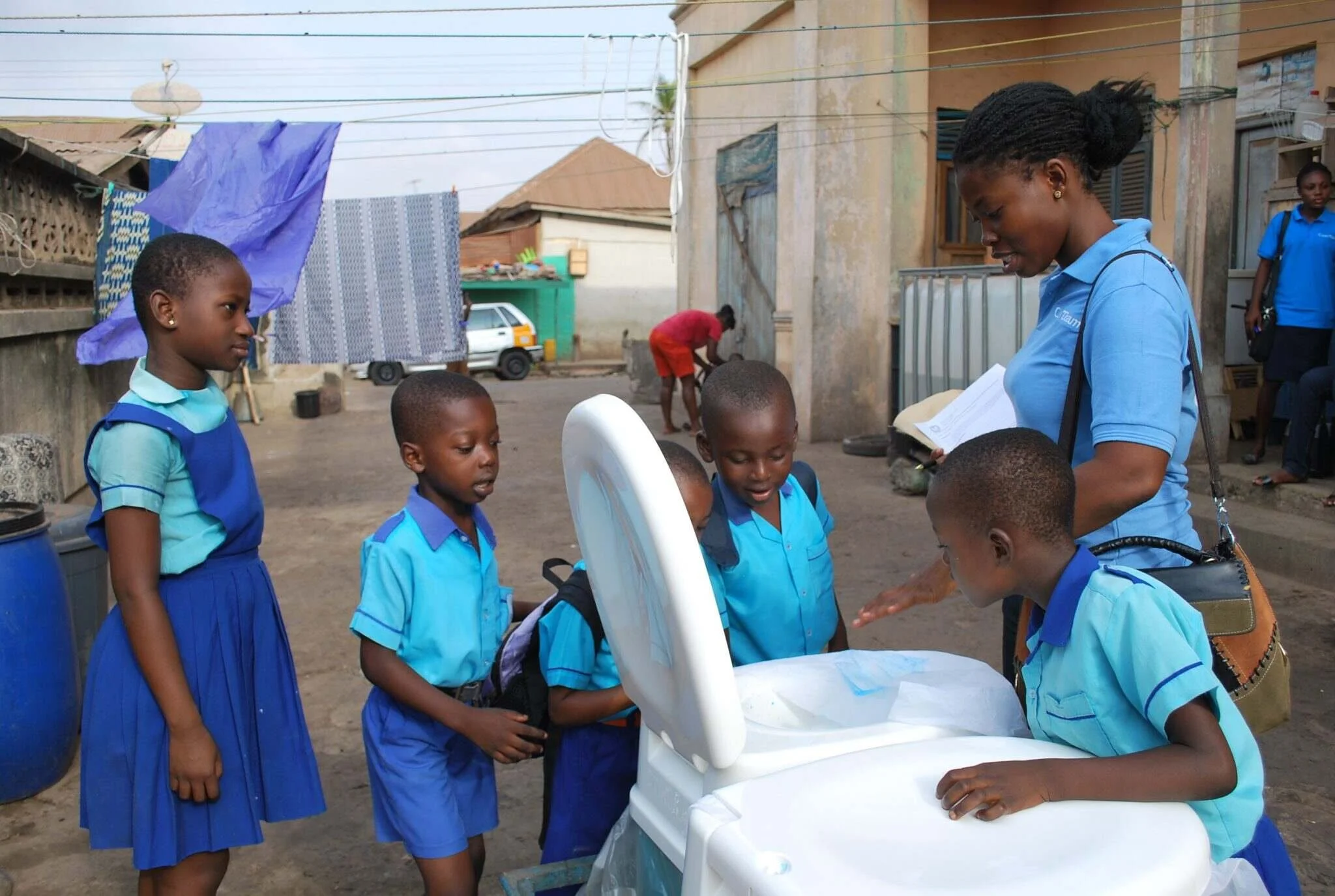 A woman in a blue shirt showing children how to use a white washing machine outdoors in a neighborhood, with houses and a person cleaning in the background.