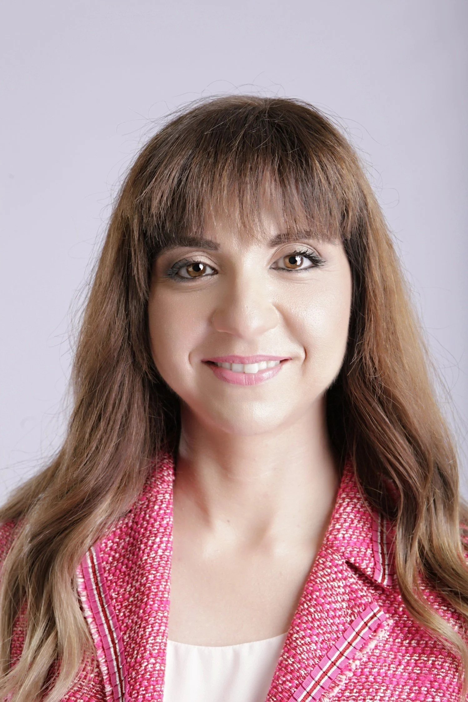 A woman with brown hair and brown eyes smiling at the camera, wearing a pink and red textured blazer over a white top, with a light gray background.