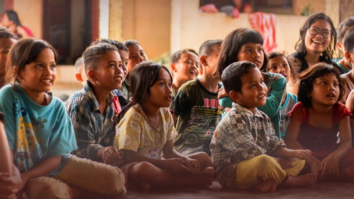 Children sitting on the floor, watching and smiling during a performance or presentation in a classroom or community setting.