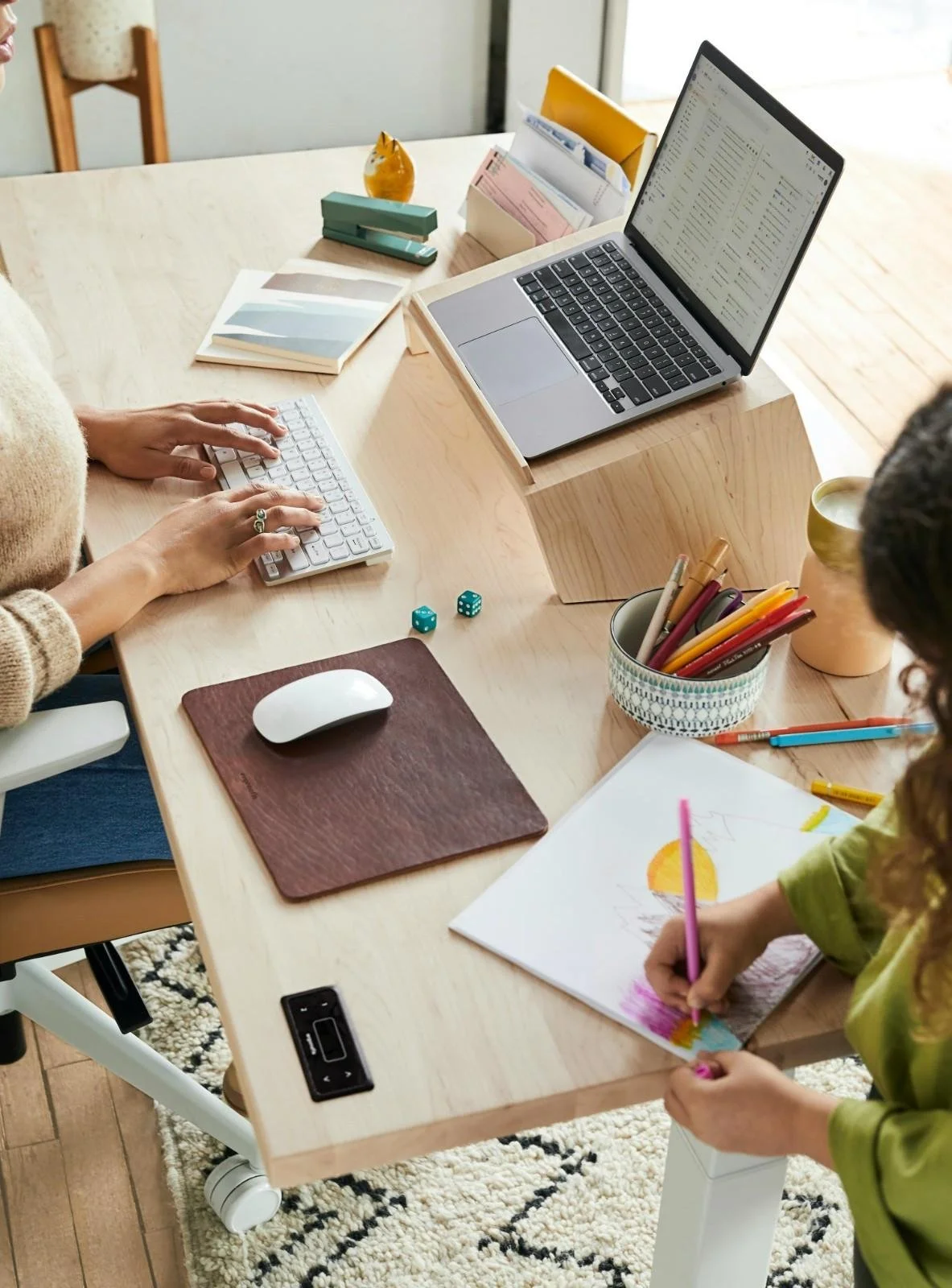 A workspace with a laptop, a keyboard, a mouse, and various office supplies on a wooden desk. Two people are working; one is typing on the keyboard, and the other is drawing with a pink crayon.