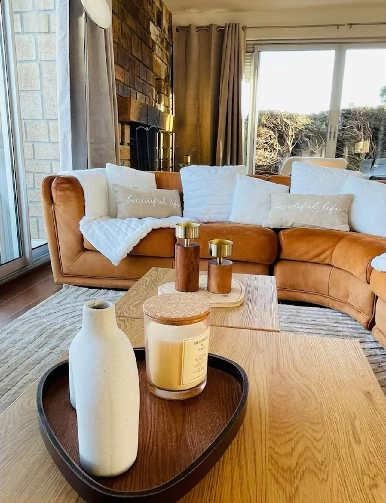 Salon avec canapé en cuir marron, coussins blancs et coussin 'beautiful life', table en bois avec bougie, vases en céramique, salle lumineuse avec grande baie vitrée et vue sur un jardin