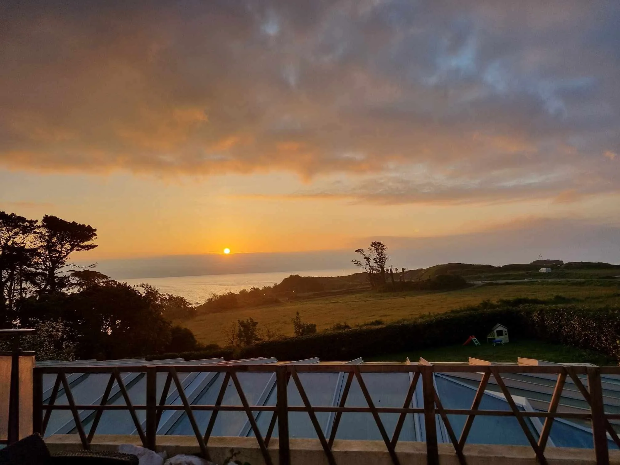 Coucher de soleil sur la mer avec des nuages gris et un paysage de collines vertes, vu depuis une terrasse avec un garde-corps en bois.