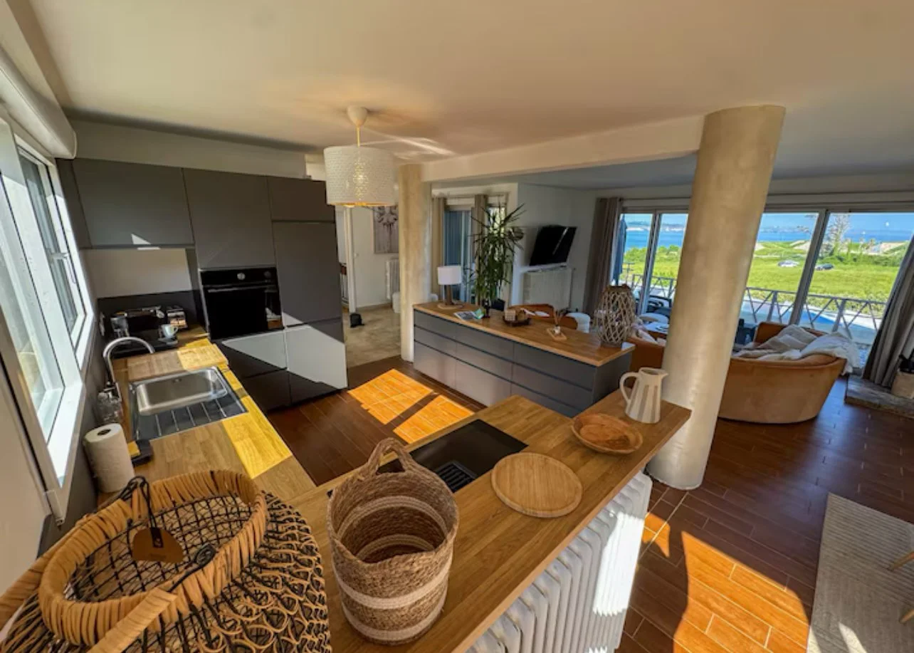 Vue de l'intérieur d'un appartement avec cuisine ouverte, salon et vue panoramique sur la mer par de grandes fenêtres.