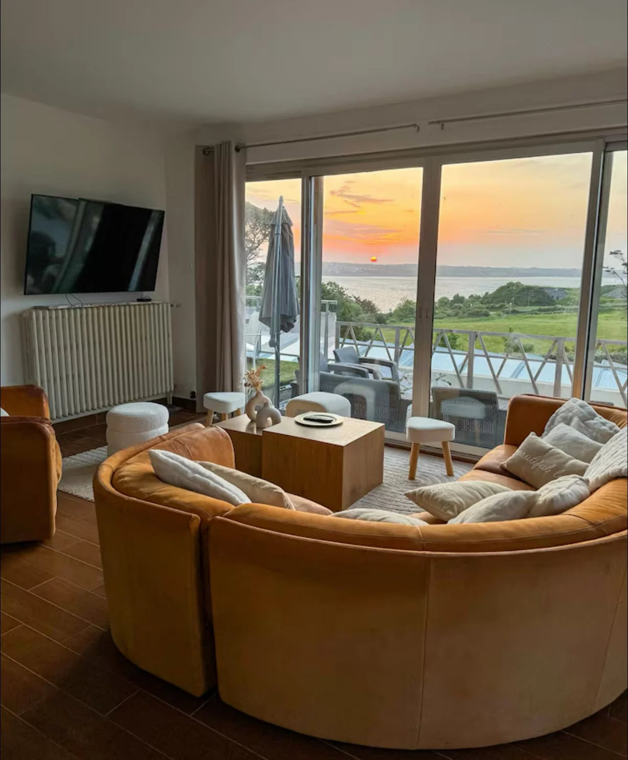 Salon moderne avec un canapé orange et des coussins blancs, face à une grande baie vitrée qui donne sur un balcon avec vue sur un lac au coucher du soleil.