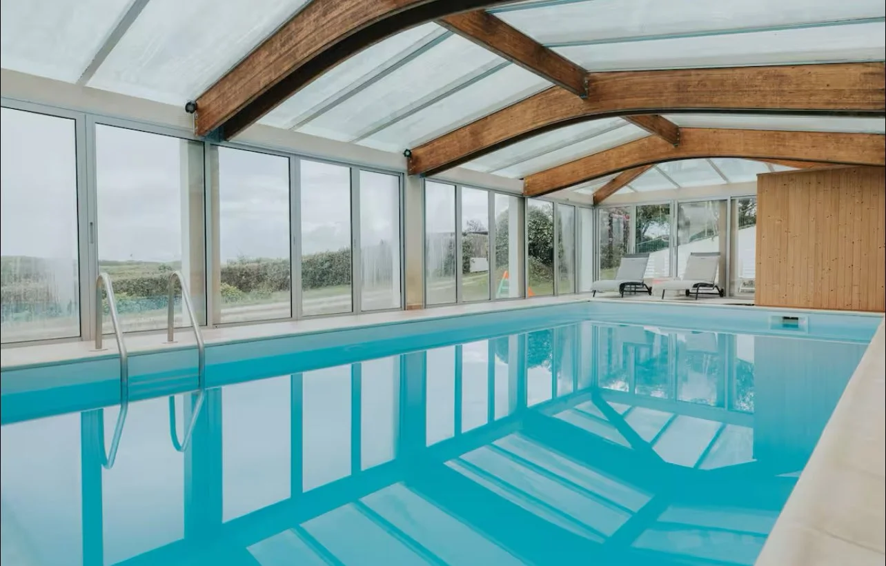 Piscine intérieure avec fenêtres panoramiques, fauteuils et structure en bois dans une ambiance lumineuse.