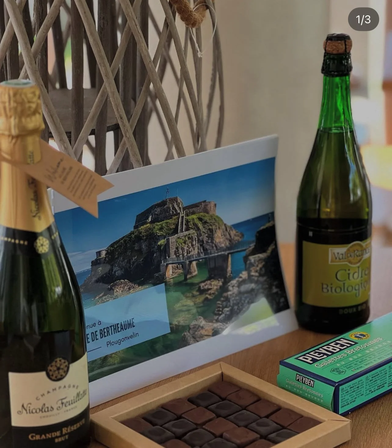 Une bouteille de champagne Nicolas Feuillatte, une bouteille de cidre biologique, une boîte de galettes bretonnes Pleyben, un coffret de chocolates et une photo d'un château sur un tableau.