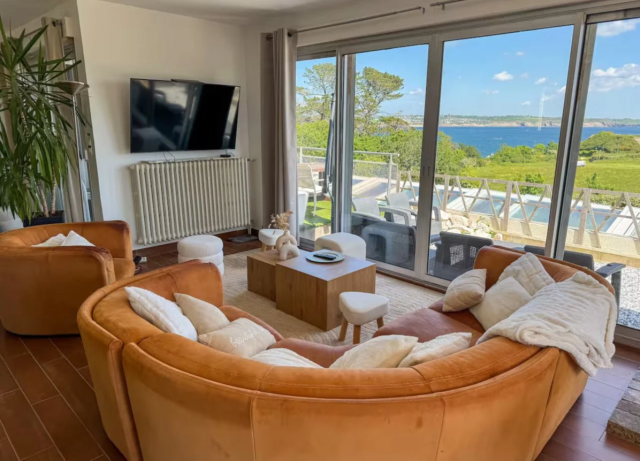 Salon avec canapés en cuir orange, vue sur un paysage côtier avec mer, arbres et ciel bleu à travers de grandes fenêtres.