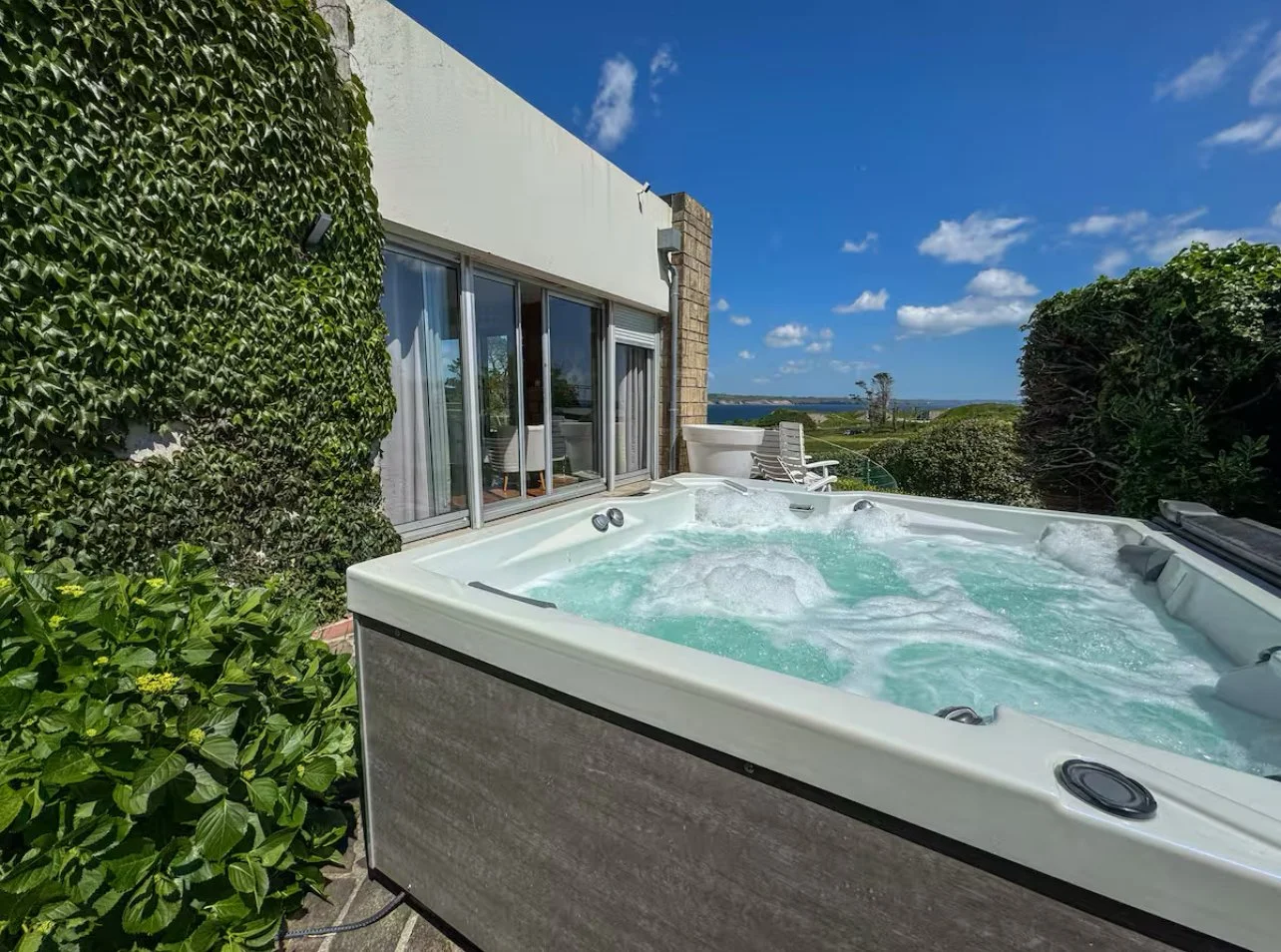 Jacuzzi extérieur chaud avec vue sur un paysage verdoyant et un ciel bleu avec quelques nuages, partiellement entouré de végétation et près d'une maison avec des fenêtres vitrées.