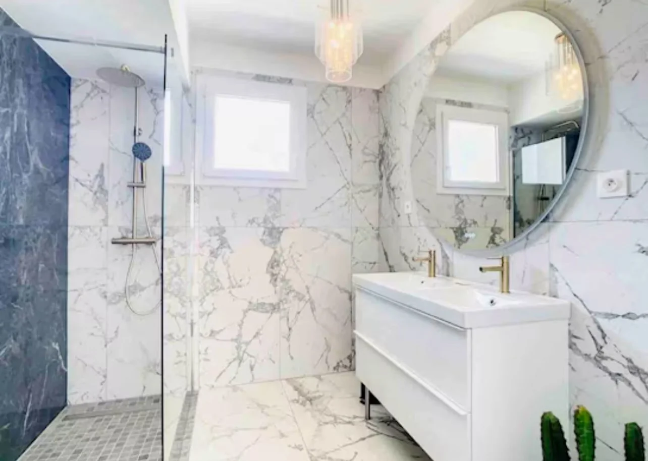 Salle de bain moderne avec murs en marbre blanc à veines grises, douche à l'italienne avec une paroi en verre, double lavabo blanc avec robinets en or, grand miroir rond, deux fenêtres pour la lumière naturelle, plante succulente verte sur le sol.