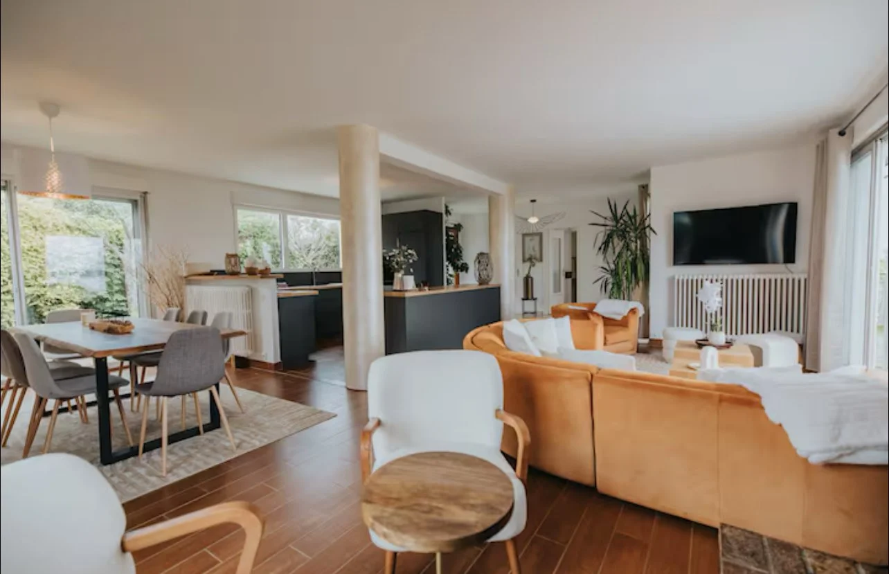 Salon moderne avec canapés en cuir beige, table basse, télévision à écran plat, espace repas avec table et chaises grises, grandes fenêtres laissant entrer la lumière naturelle, décoration végétale et lumineuse.