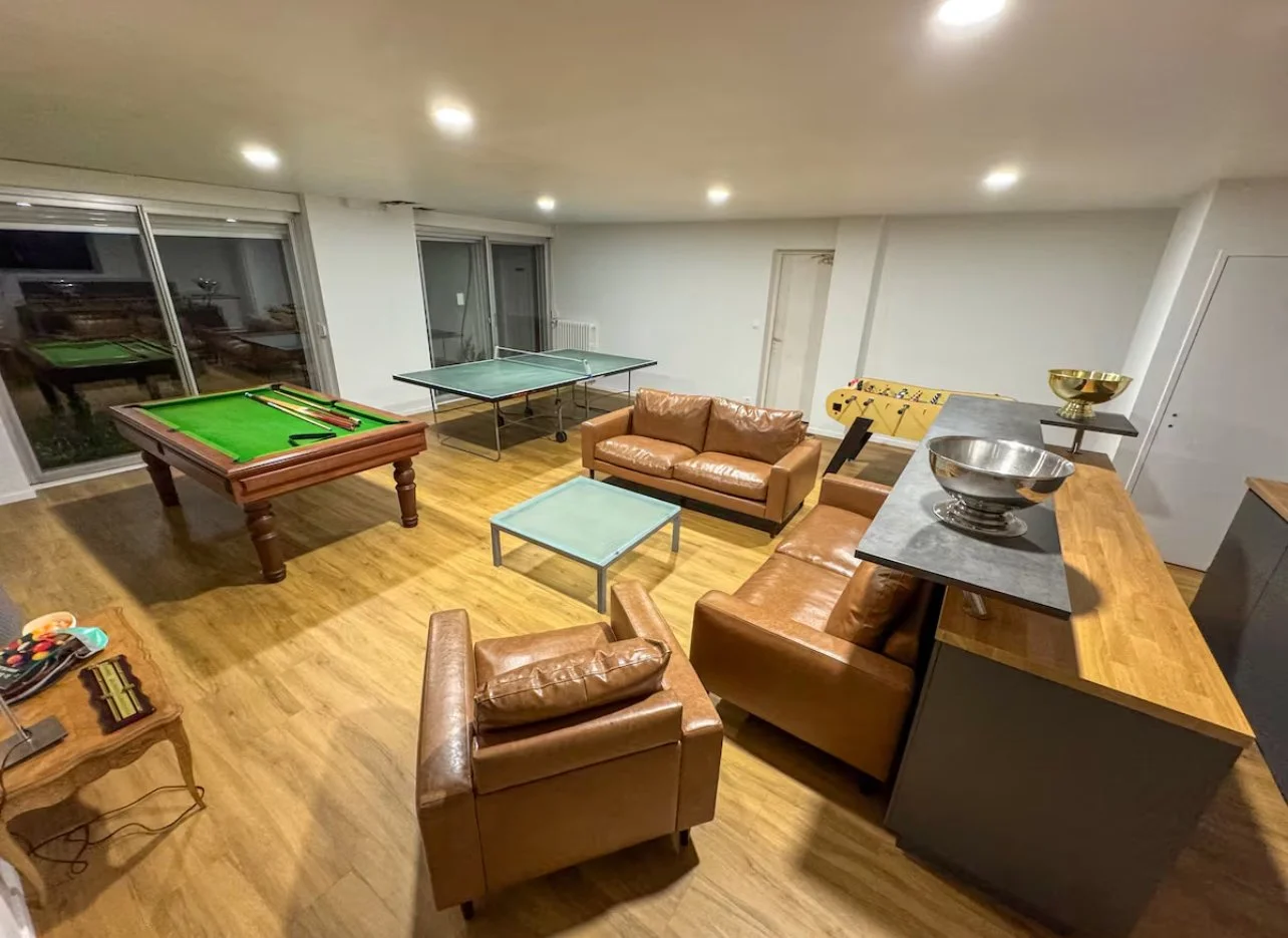 Salle de jeux avec table de billard, table de ping-pong, canapés en cuir marron, table basse, et bureau avec comptoir à boissons, dans une pièce lumineuse.