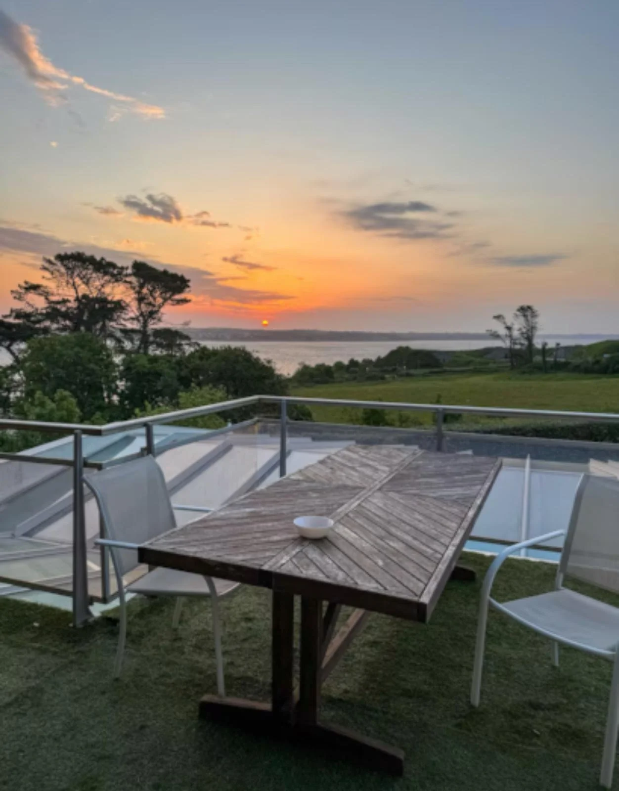 Vue d'un coucher de soleil derrière un lac avec un ciel clair et quelques nuages, vue depuis une terrasse avec une table en bois et des chaises blanche.