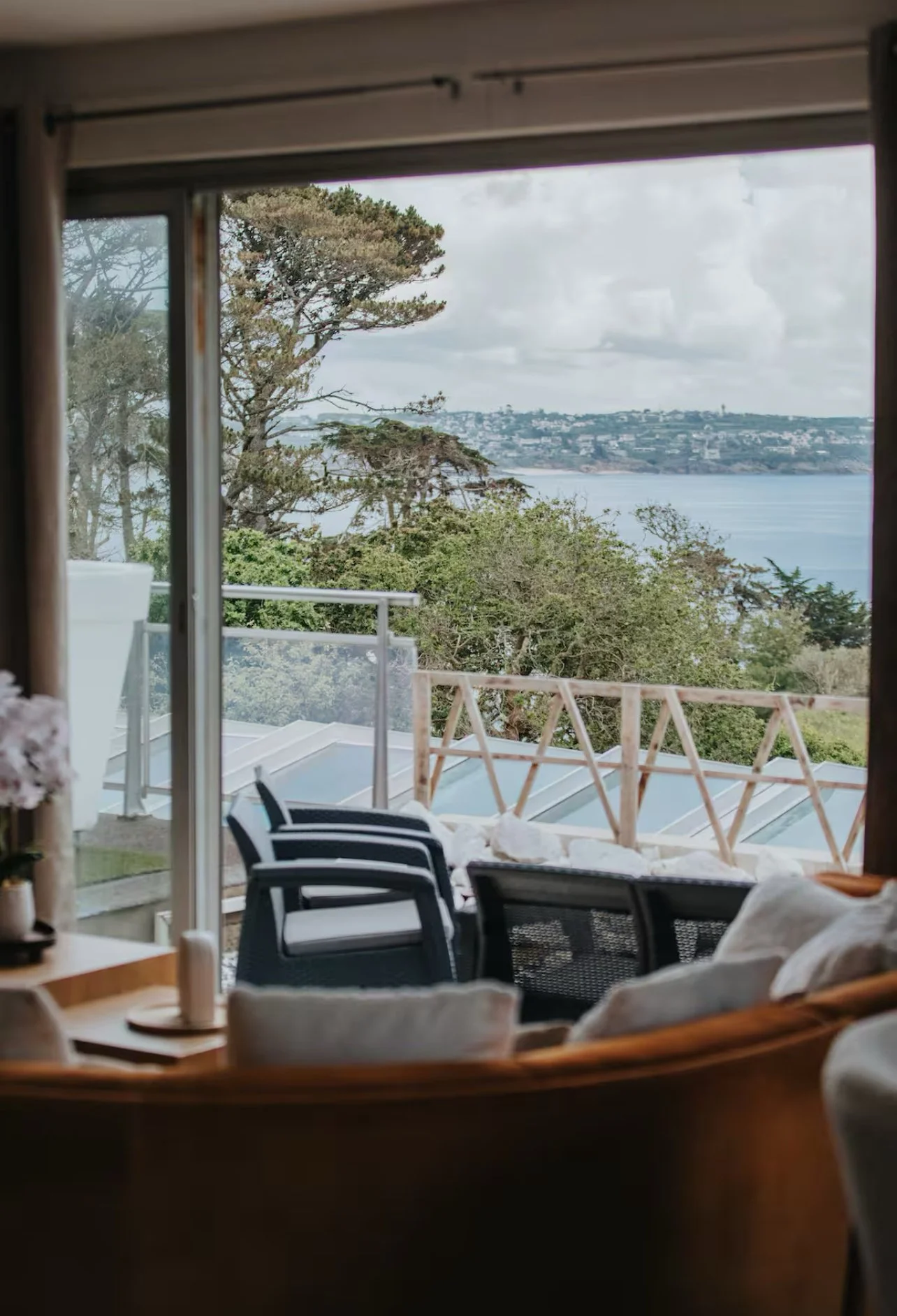 Vue depuis une pièce intérieure vers un balcon avec des chaises en plastique et un mobilier en bois, regardant vers des arbres et une étendue d'eau sous un ciel nuageux.